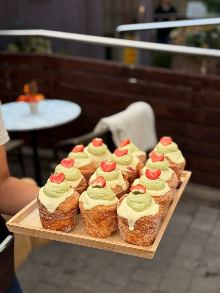 Tray of twelve baked cupcakes topped with green frosting and small heart-shaped red candies, held by a person outdoors.