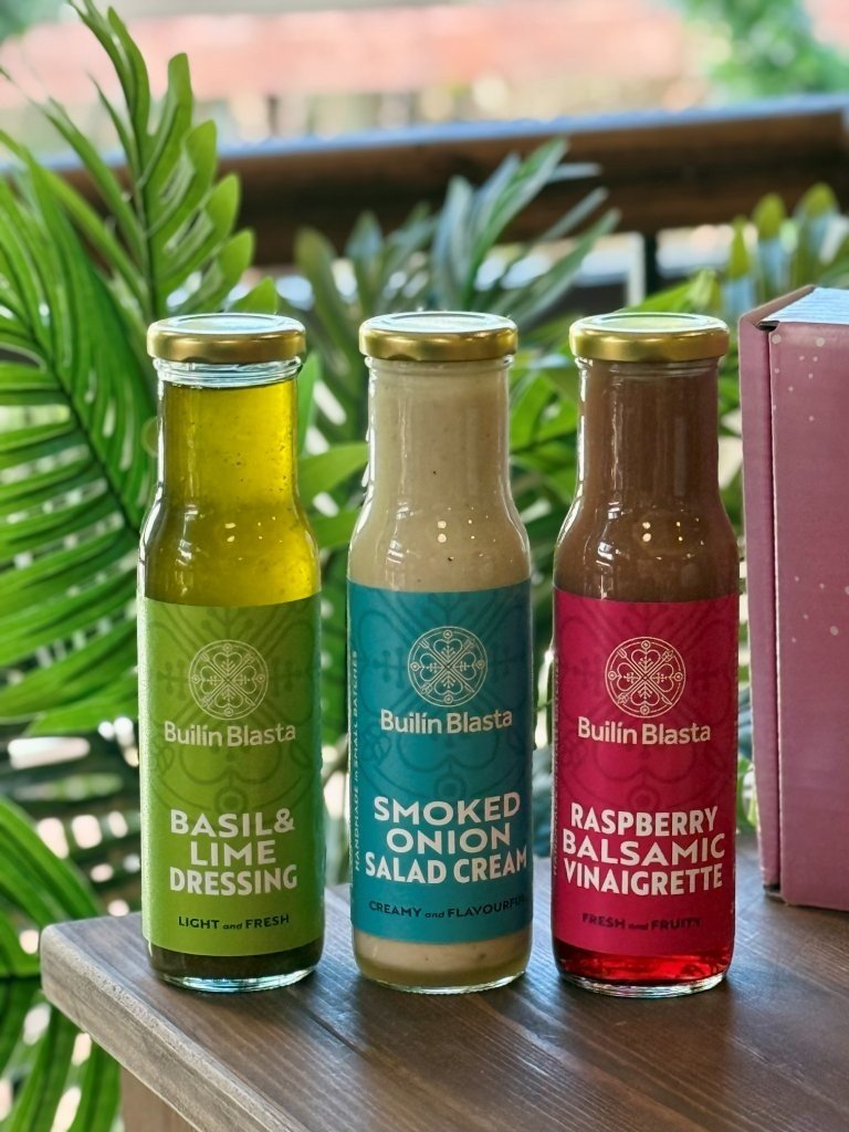 Three bottled salad dressings on a wooden surface, with green plants and a blurred outdoor background. The dressings are basil & lime, smoked onion salad cream, and raspberry balsamic vinaigrette.