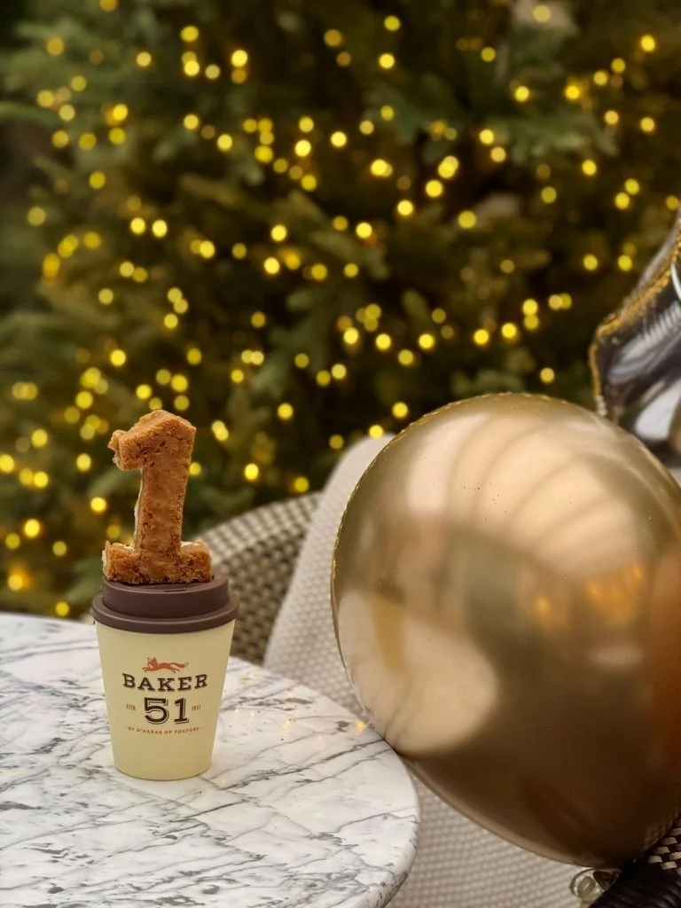 A cake topper in the shape of the number 1 on a small cake or dessert cup, with a large gold balloon and blurred Christmas tree with yellow lights in the background.