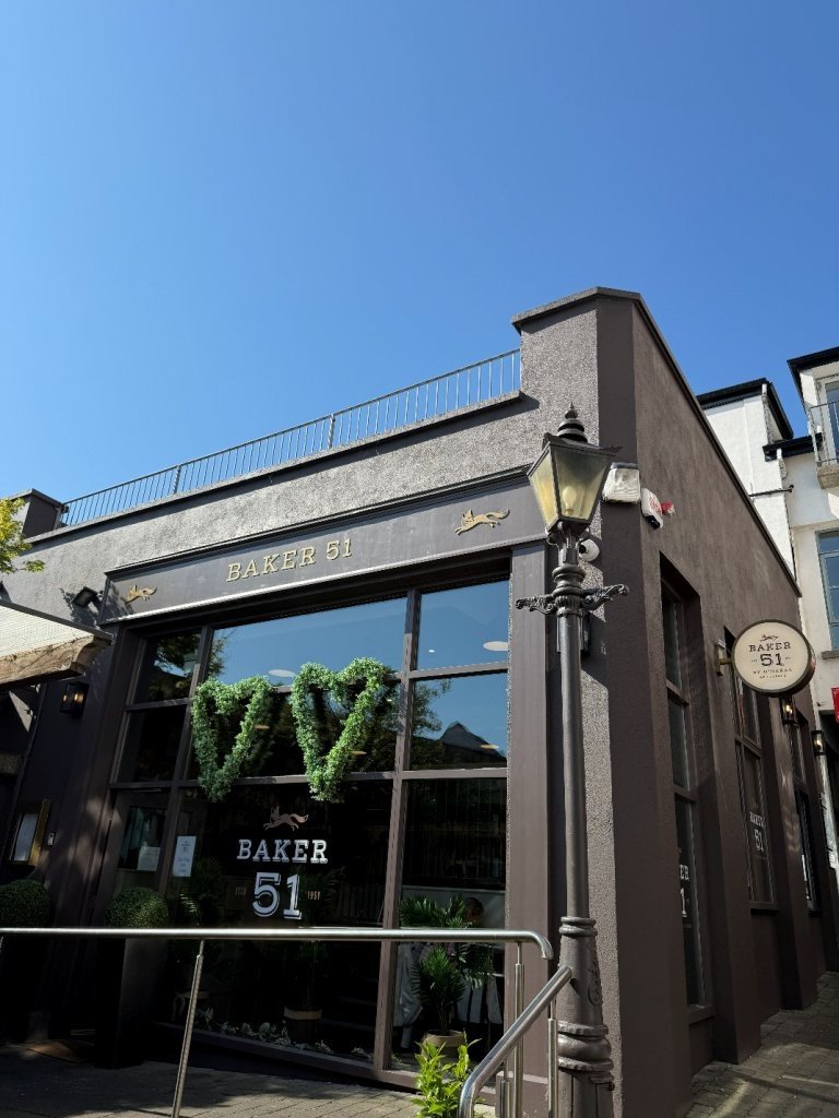 Exterior view of a gray building with large glass windows, a sign reading 'BAKER 51', and decorative fish symbols above the sign. A window display with a green heart-shaped decoration inside.