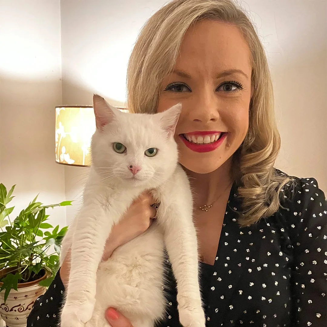 Person smiling and holding a white cat indoors next to a potted plant.