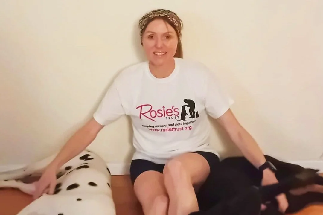Woman sitting between a Dalmatian and a black dog, wearing a T-shirt with "Rosie's Trust" logo, indoors.