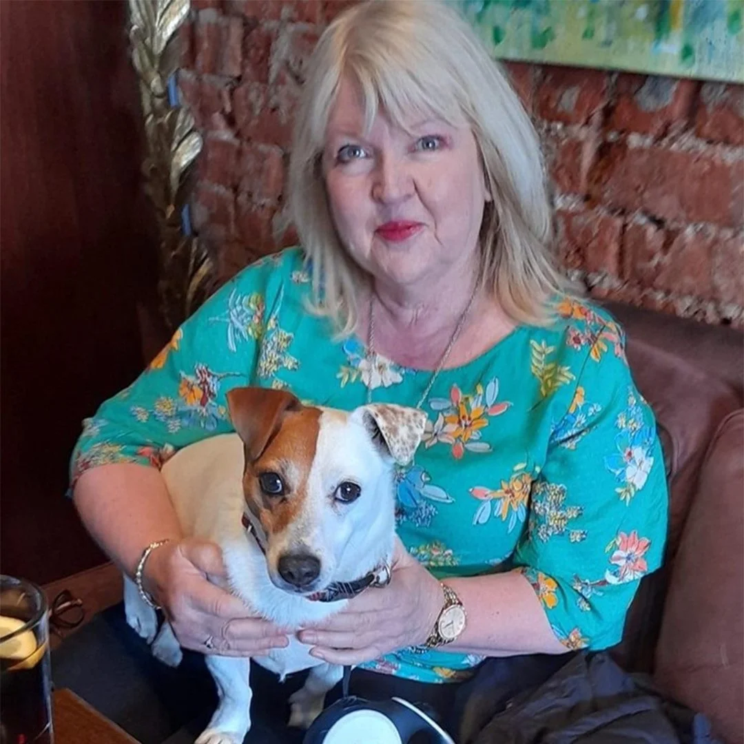 A woman with blonde hair sitting in a cafe, holding a small brown and white dog. The woman is wearing a floral green blouse. The background features a rustic brick wall.