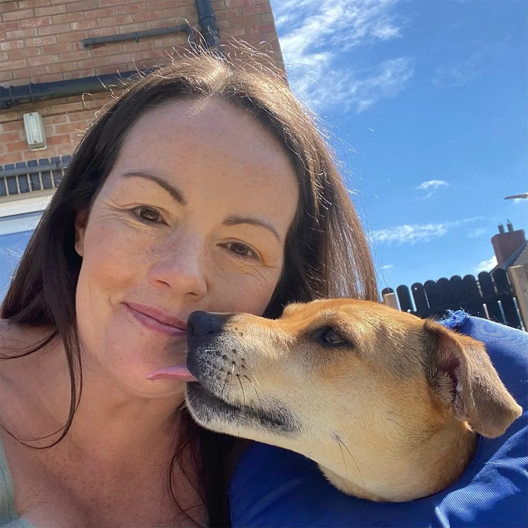 Woman with long brown hair smiling and a dog with brown fur licking her face, wearing a blue protective collar, outdoors on a sunny day.