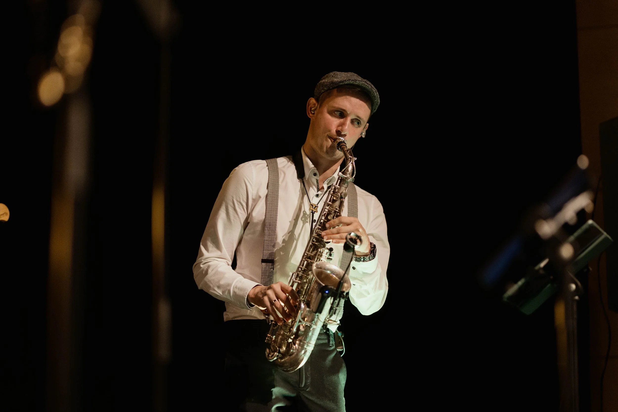 Ein junger Mann spielt Saxophon in einem dunklen Raum, trägt eine weiße Hemd, eine Weste, eine Mütze und hat Kopfhörer auf, während er Musik macht.