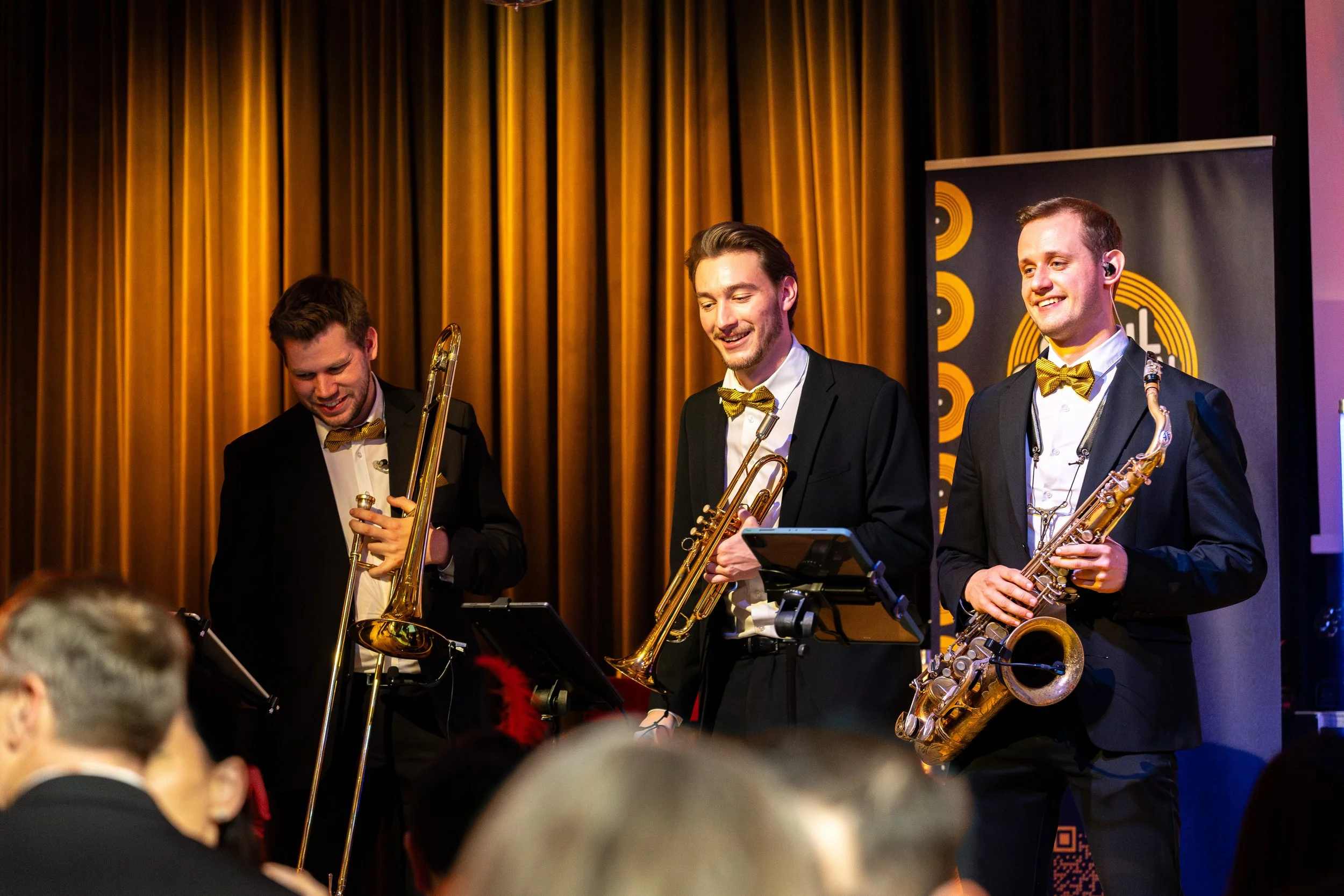 Drei Musiker in schwarzen Anzügen mit gelben Fliege spielen Saxophon bei einem Konzert vor einem goldenen Vorhang.