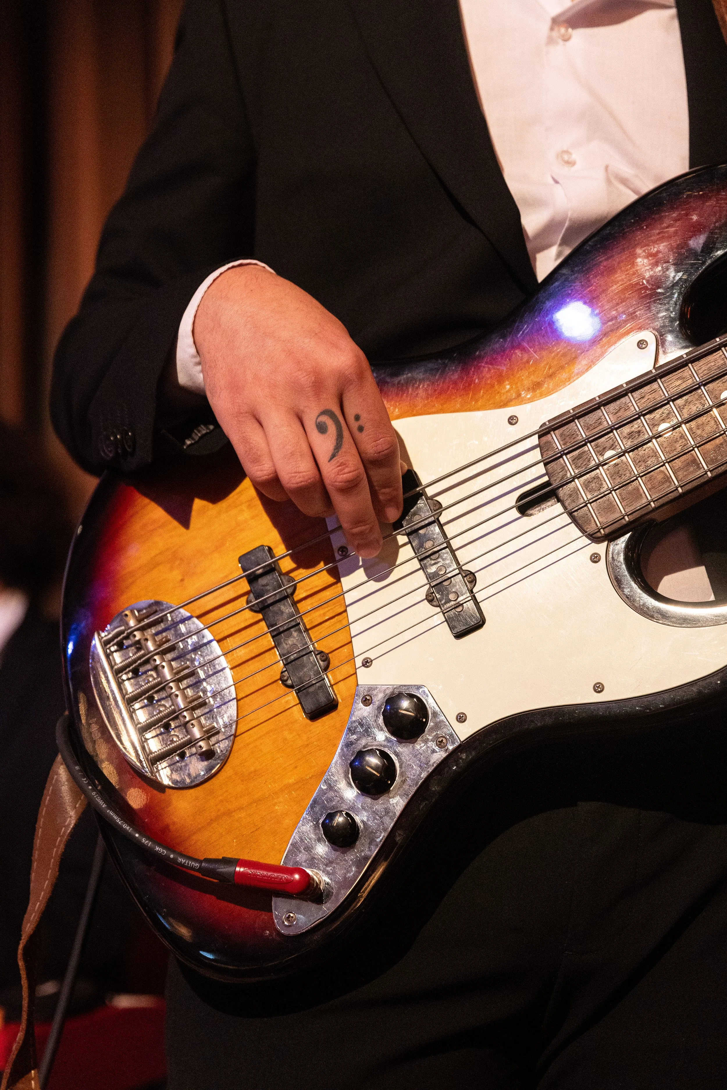 Person in formeller Kleidung, die eine elektrische Gitarre spielt, sichtbar ist nur der obere Teil des Körpers und die Gitarre, mit Fokus auf die Hand, die die Saiten hält und die Steuerung. Die Person trägt einen schwarzen Anzug und ein weißes Hemd.