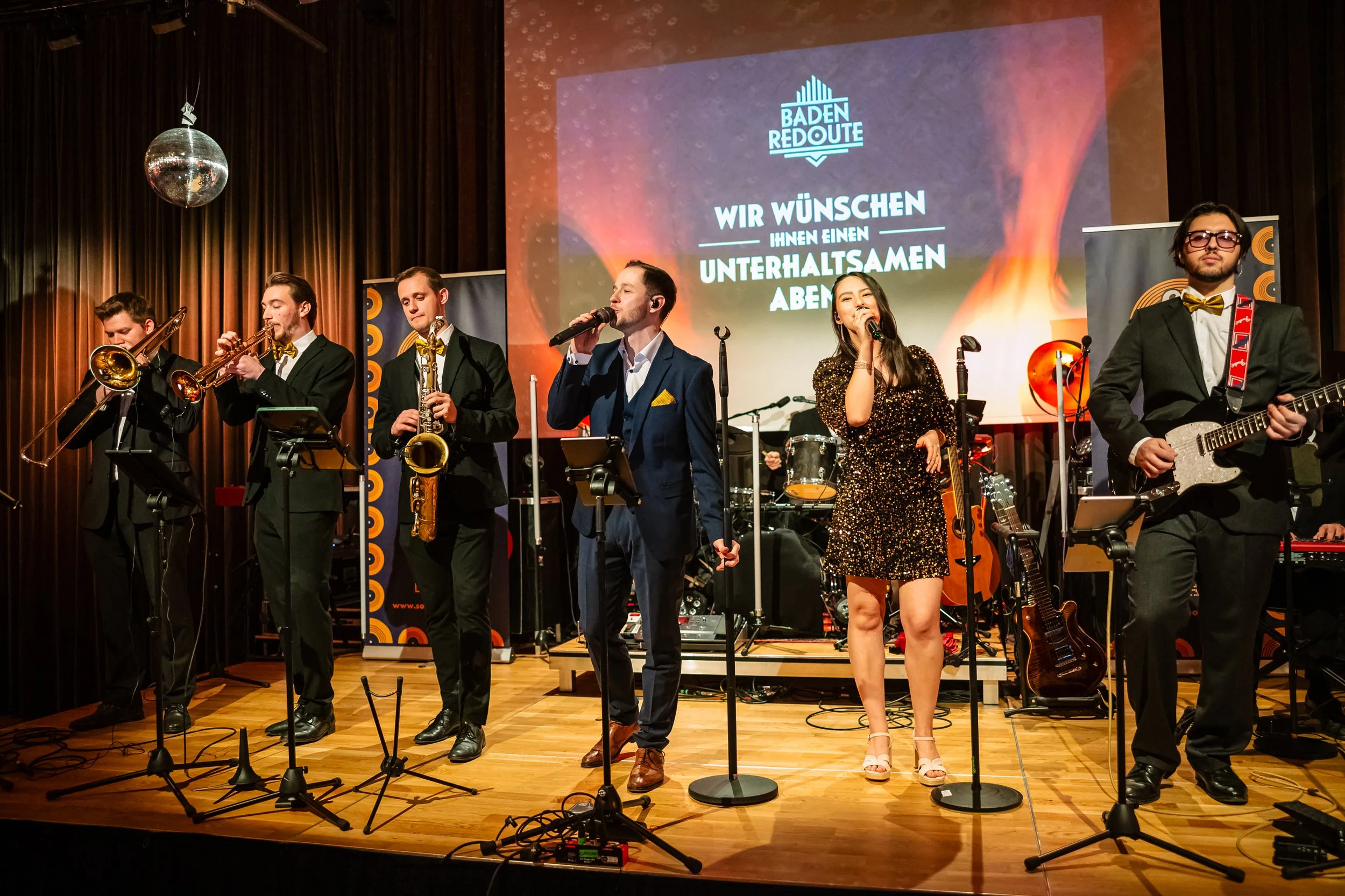 Eine Band aus sechs Musikern spielt auf einer Bühne mit dunklen Vorhängen im Hintergrund. Zwei Sängerinnen, drei Männer mit Blasinstrumenten und einer mit E-Gitarre. Eine große Leinwand im Hintergrund zeigt das Logo 'Baden Redoute' und den Text 'Wir wünschen Ihnen einen unterhaltsamen Abend'.