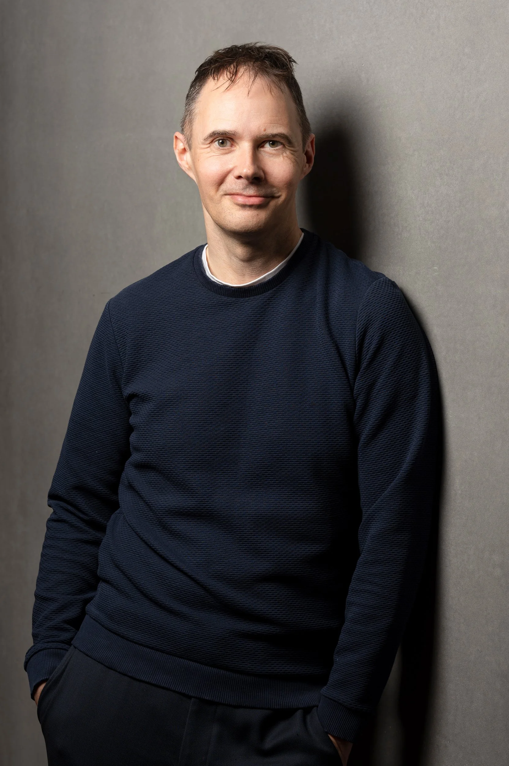 Portrait of a man with short brown hair, wearing a navy sweater, leaning against a gray wall with his hands in his pockets, smiling slightly.