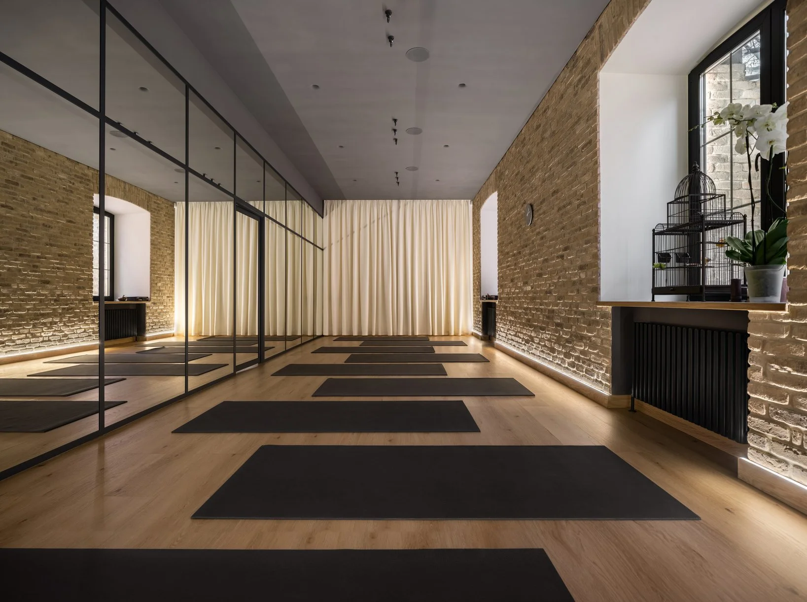 An empty yoga studio with dark mats on a warm wooden floor, exposed brick walls, and soft natural light.