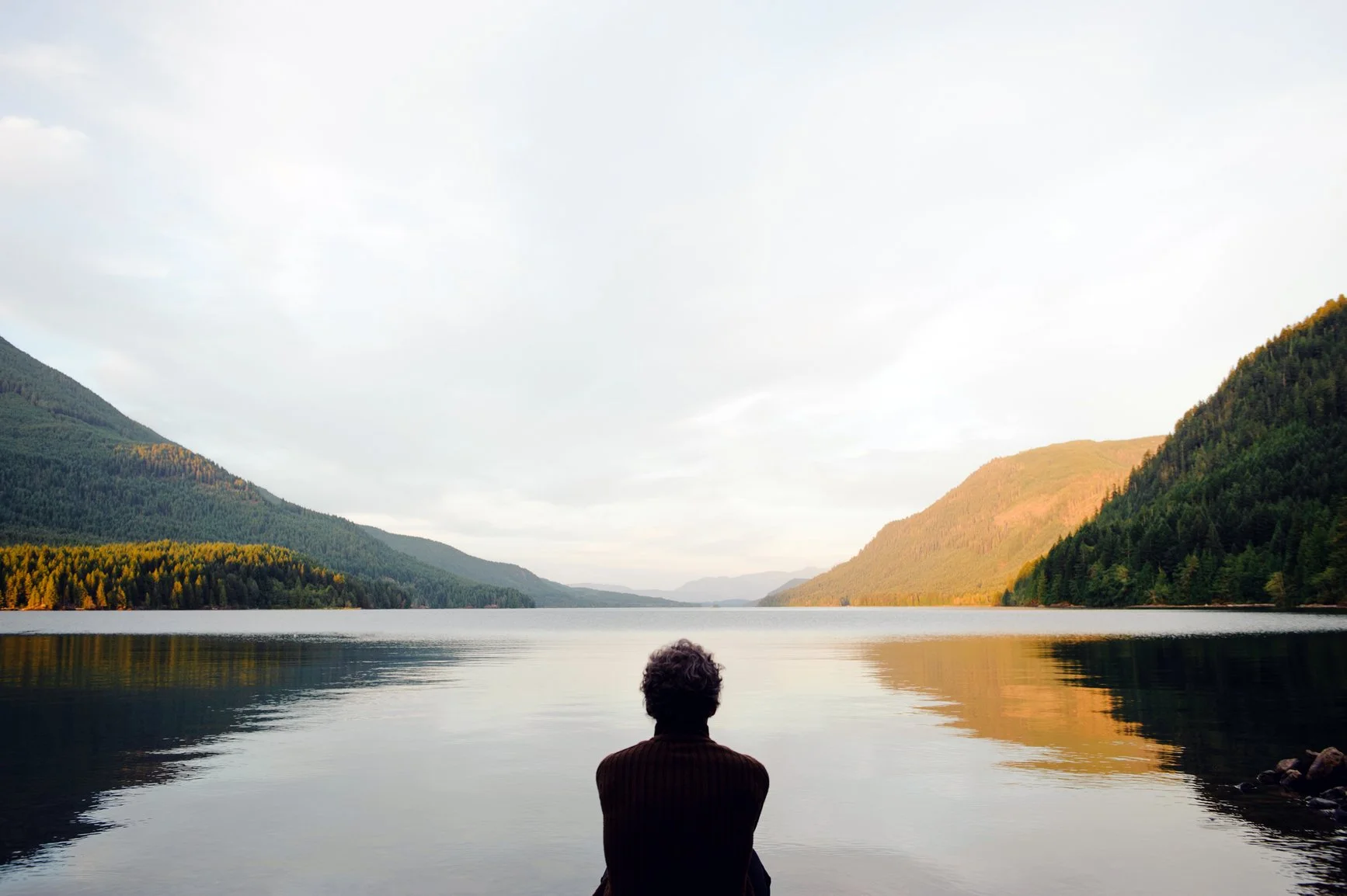 Person sitting at the edge of a still lake at dusk, looking out toward distant mountains.