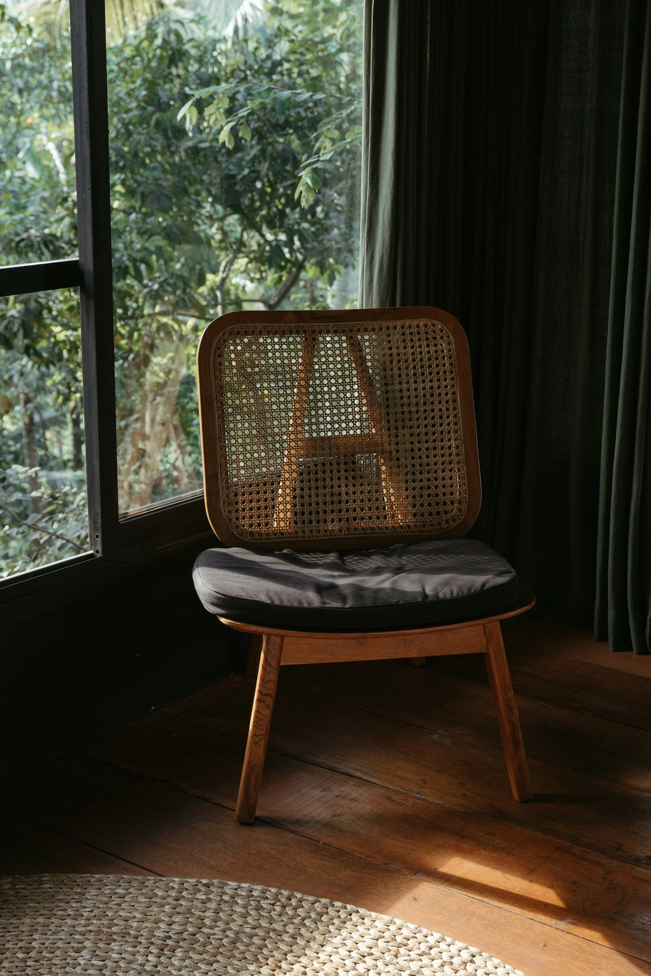 Cane-back chair with a dark cushion by a window, warm light filtering through greenery outside