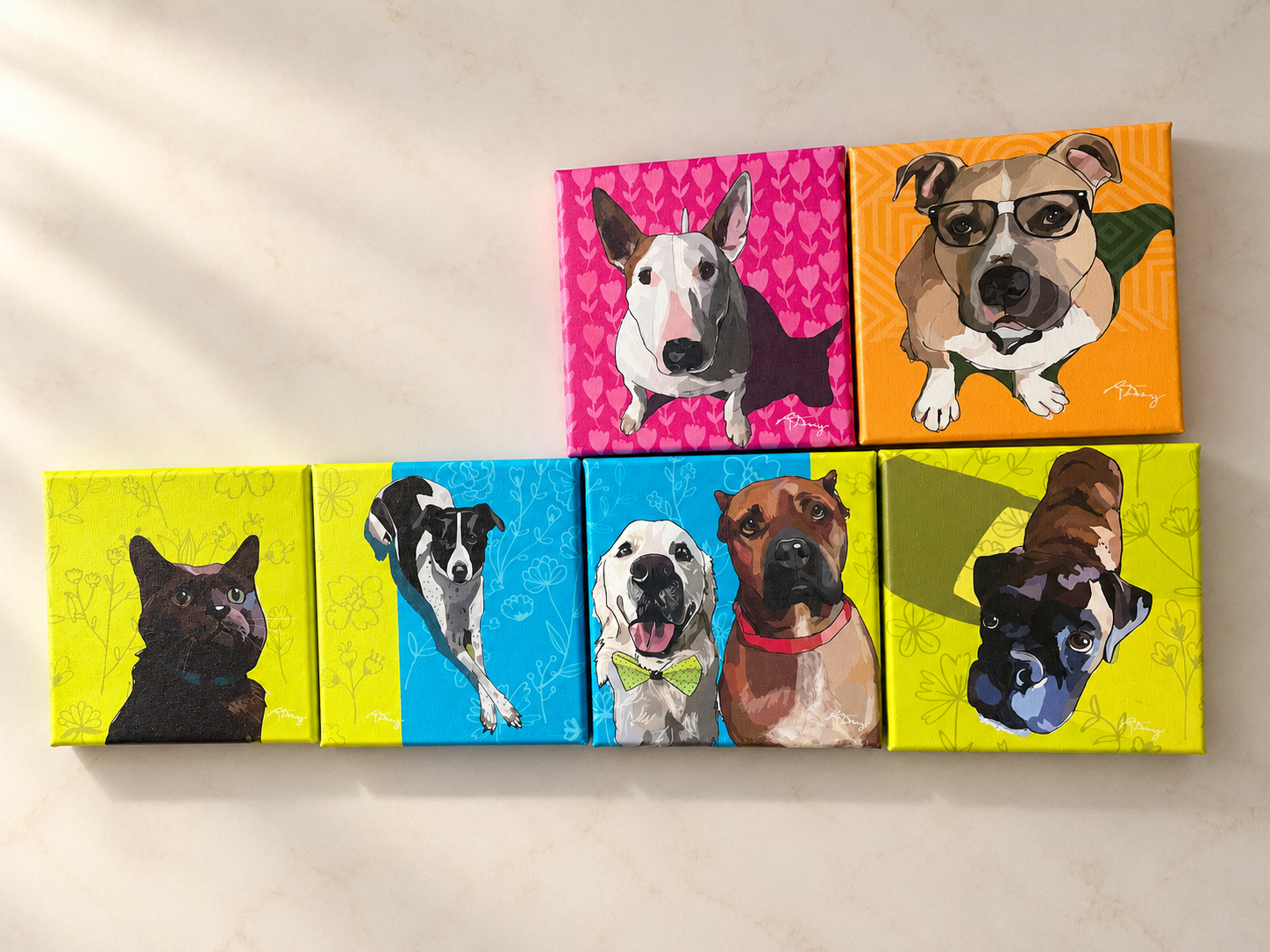 Six colorful canvases with portrait illustrations of different dogs and a cat, arranged in two rows on a light-colored surface. The paintings feature vibrant backgrounds with flower or geometric patterns.