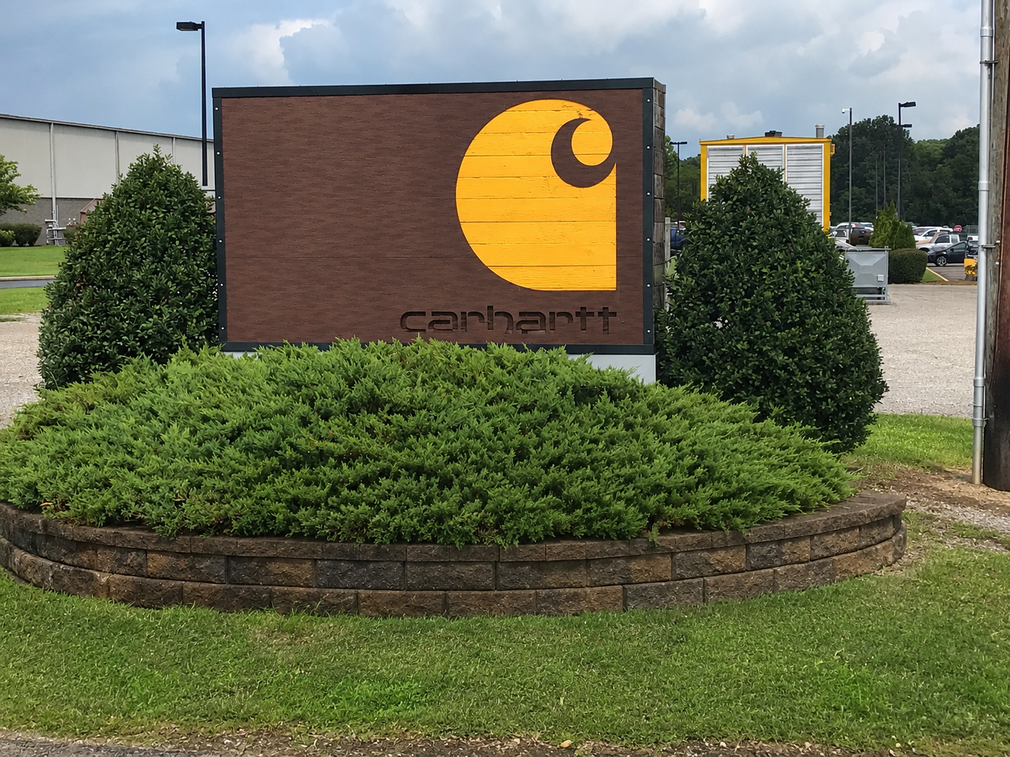 Carhartt store sign in Ohio with a yellow and black logo on a wooden background, surrounded by green bushes and plants, in a parking lot.