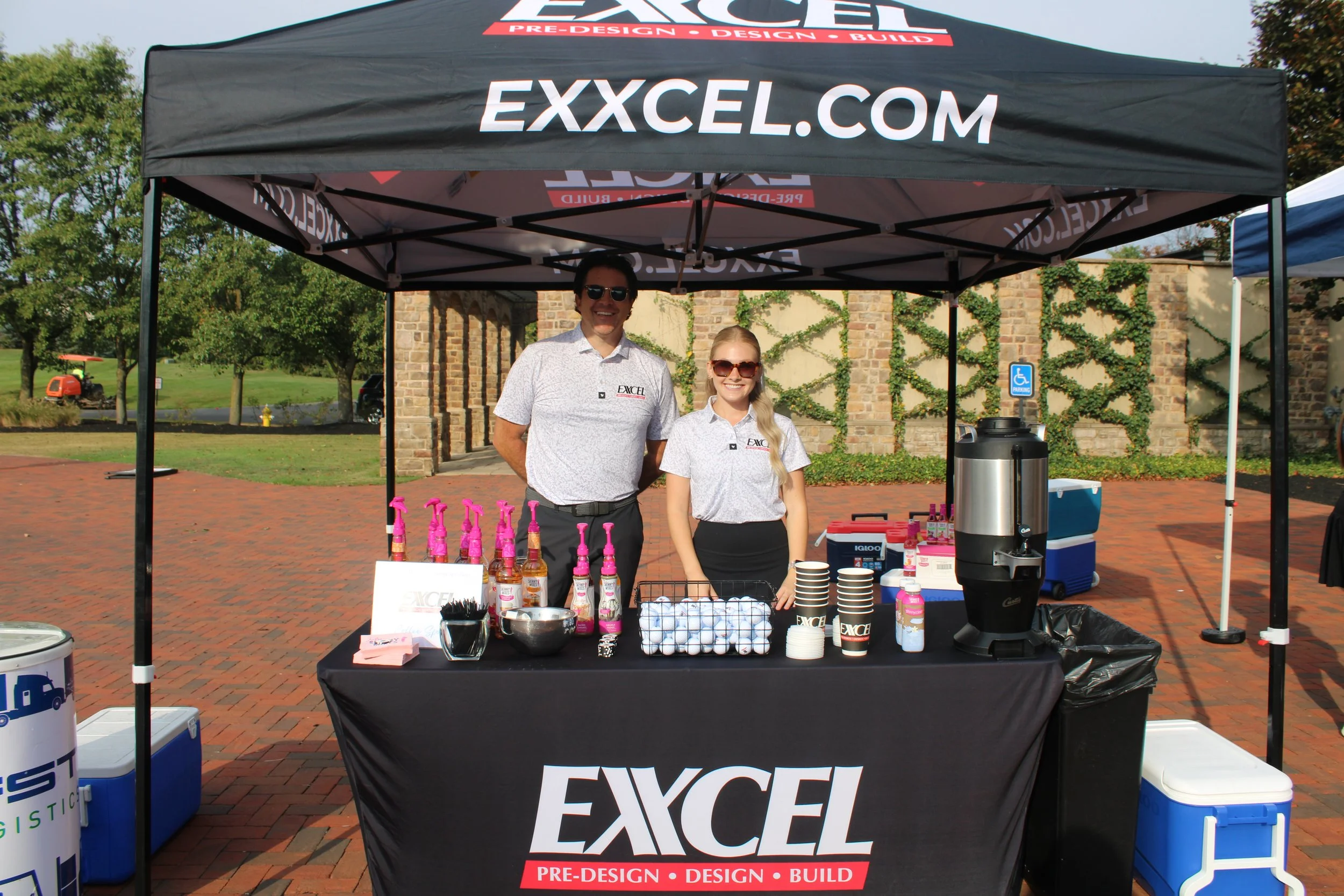 Two smiling individuals, a man and a woman, stand behind a black table in front of a black canopy tent with the website 'EXCEL.COM' and the words 'PRE-DESIGN, DESIGN, BUILD.' The table is covered with bottles of pink spray, cups, and other promotional items. The background features brick buildings, trees, and a blue sky.