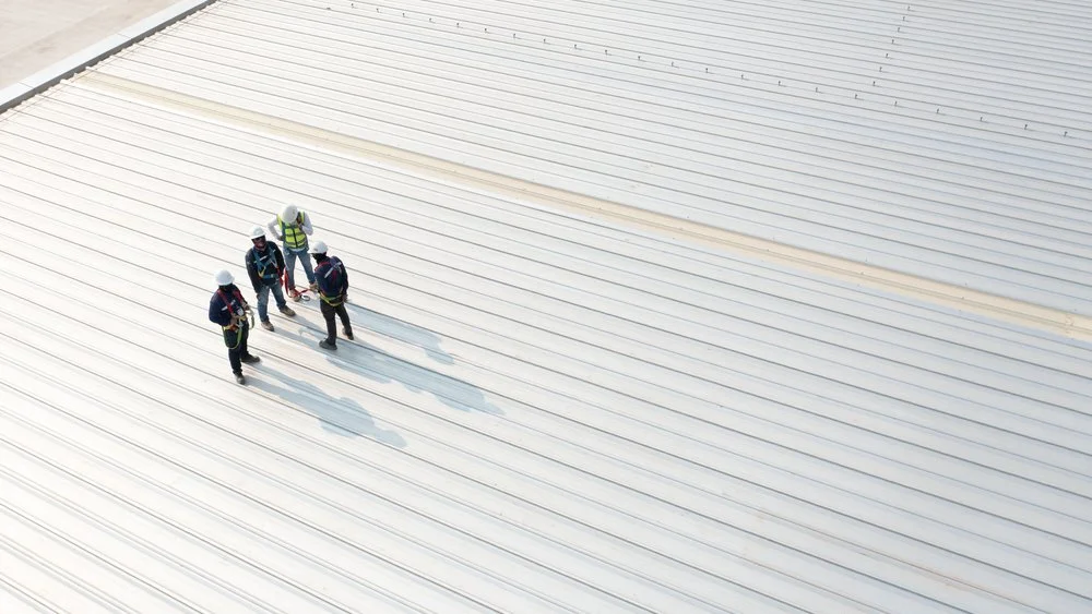 Four workers wearing helmets and safety vests standing on a large, white, metal roof on an industrial warehouse.
