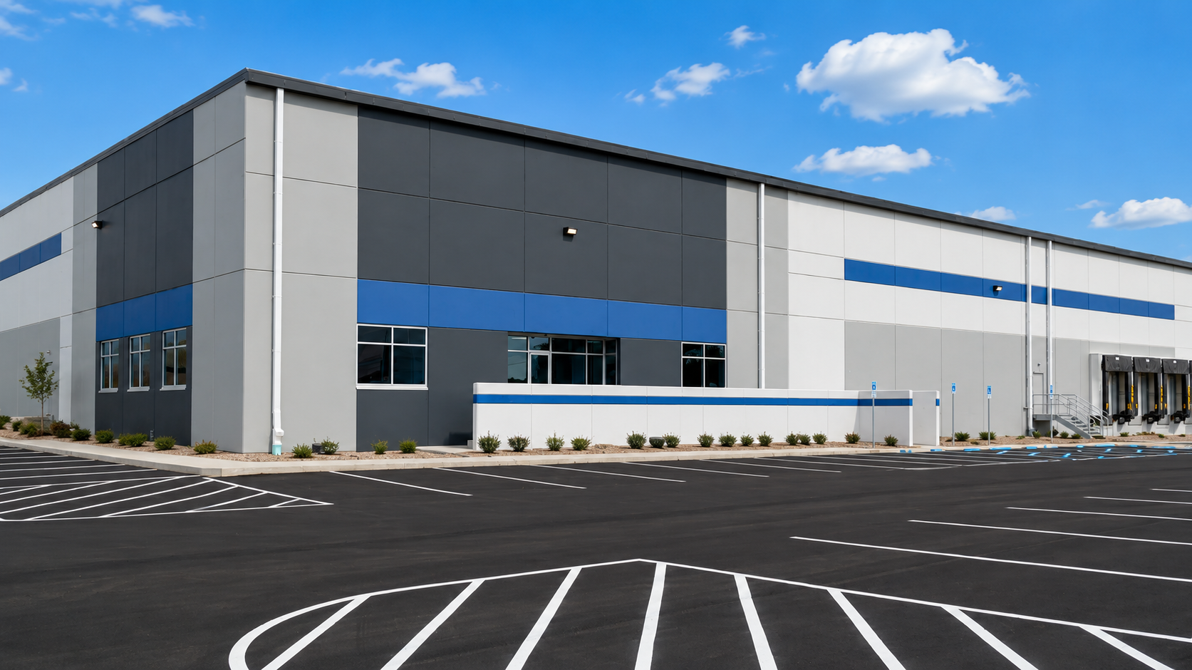 Large, modern warehouse building with gray, white, and blue panels, parking lot with striped spaces, and clear blue sky with scattered clouds.