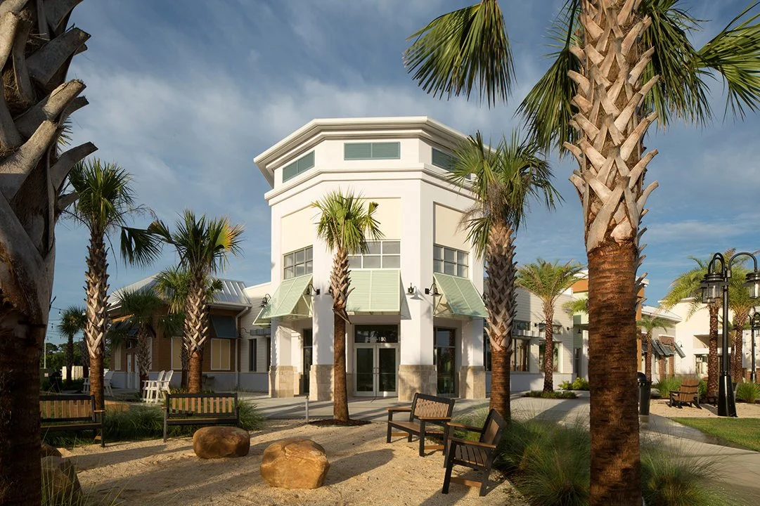 A modern building with white walls and large windows, surrounded by palm trees and outdoor benches in a landscaped area.