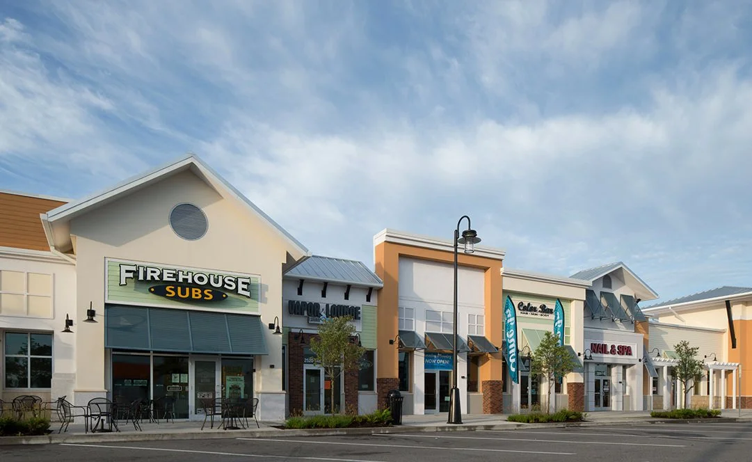 A shopping center with various retail stores, including Firehouse Subs, a nail and spa salon, and a boutique boutique, under a partly cloudy sky.