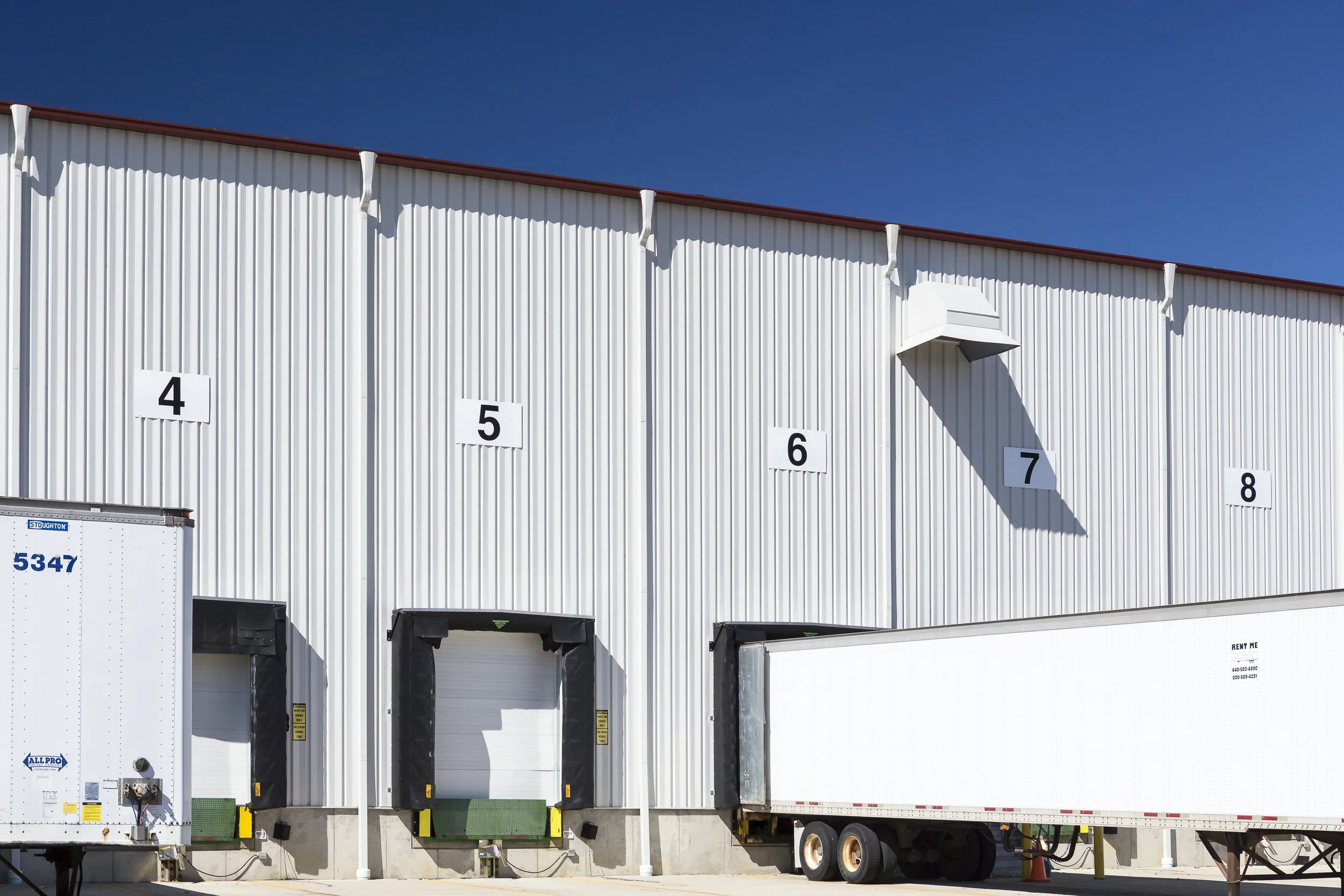 Loading docks with numbered doors on a warehouse building, with trucks parked in front and a clear blue sky above.