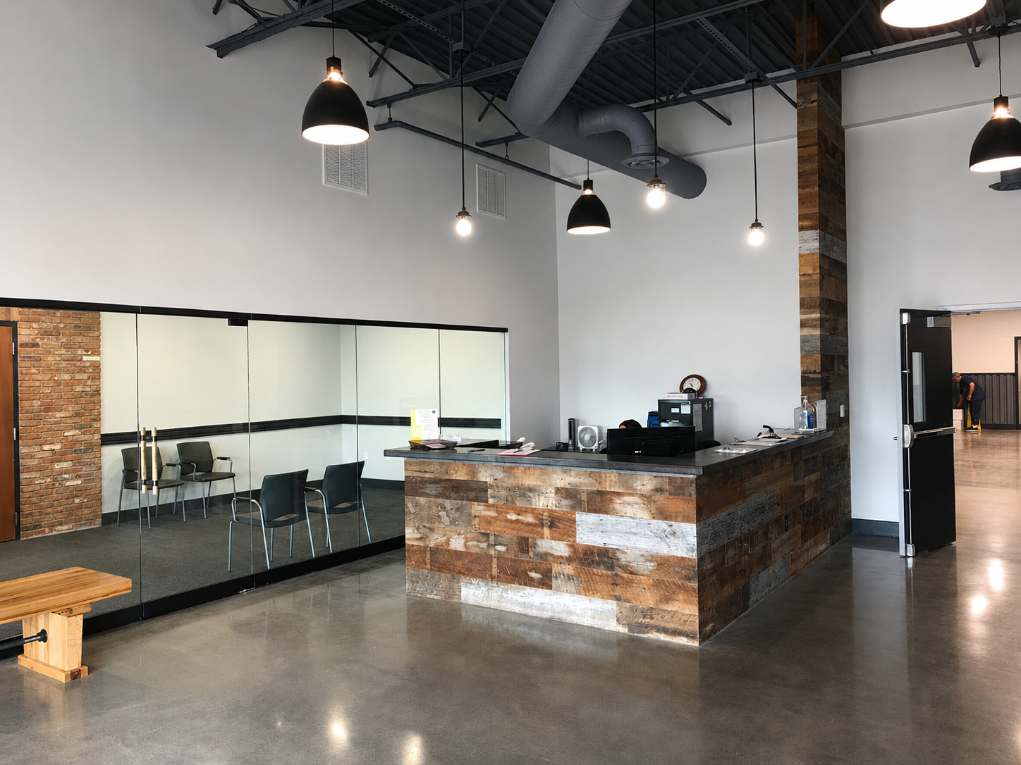 Modern reception area with a wooden front desk, a glass-walled meeting room with chairs, and industrial-style ceiling with hanging lights.