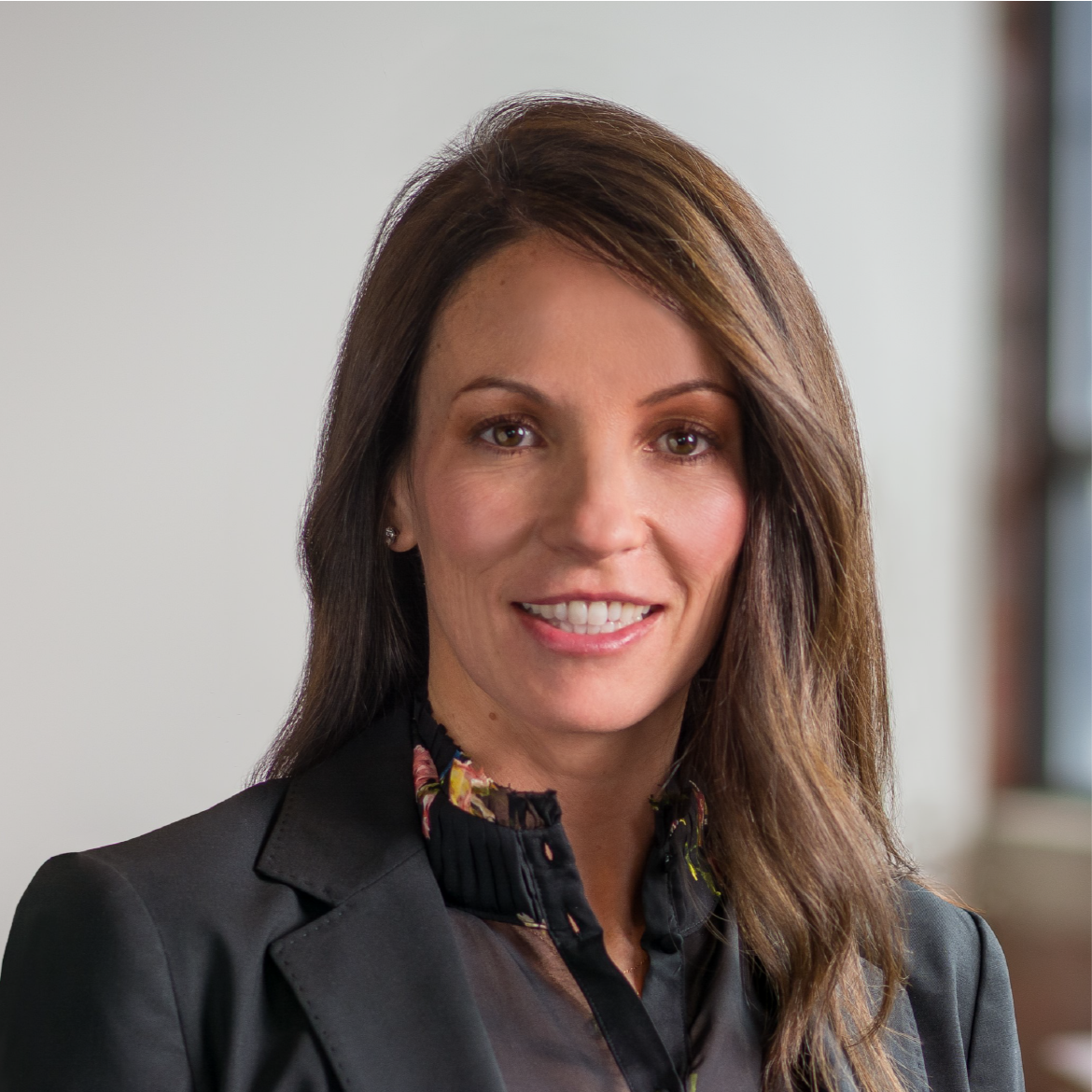Businesswoman in a black blazer and floral blouse smiling, with long brown hair, in a professional environment.