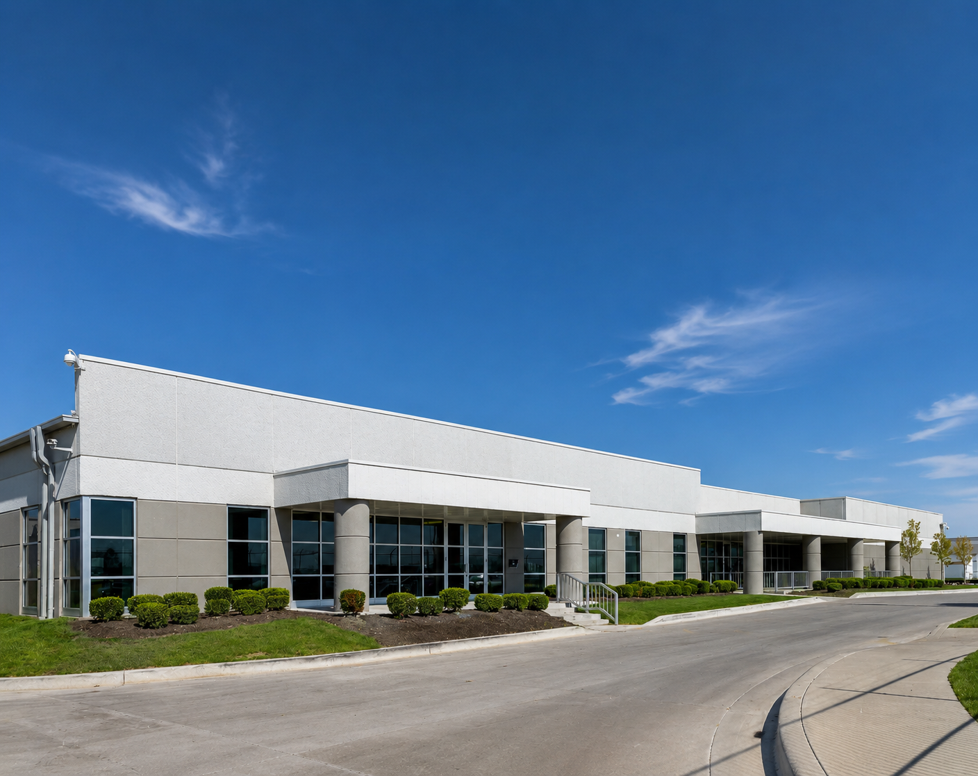 Modern commercial building with large glass windows and landscaped bushes in front, under a bright blue sky.