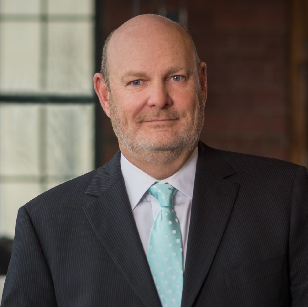 Man in a suit with a light blue tie, standing indoors with soft lighting.