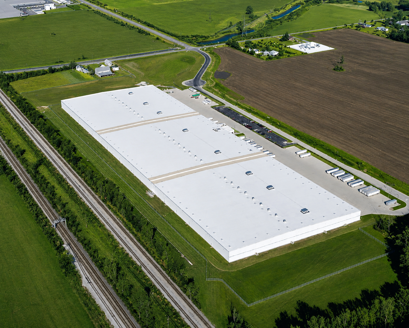 Aerial view of a large industrial warehouse with a white roof, surrounded by a parking lot with trucks and trailers, green fields, and railroad tracks on one side.