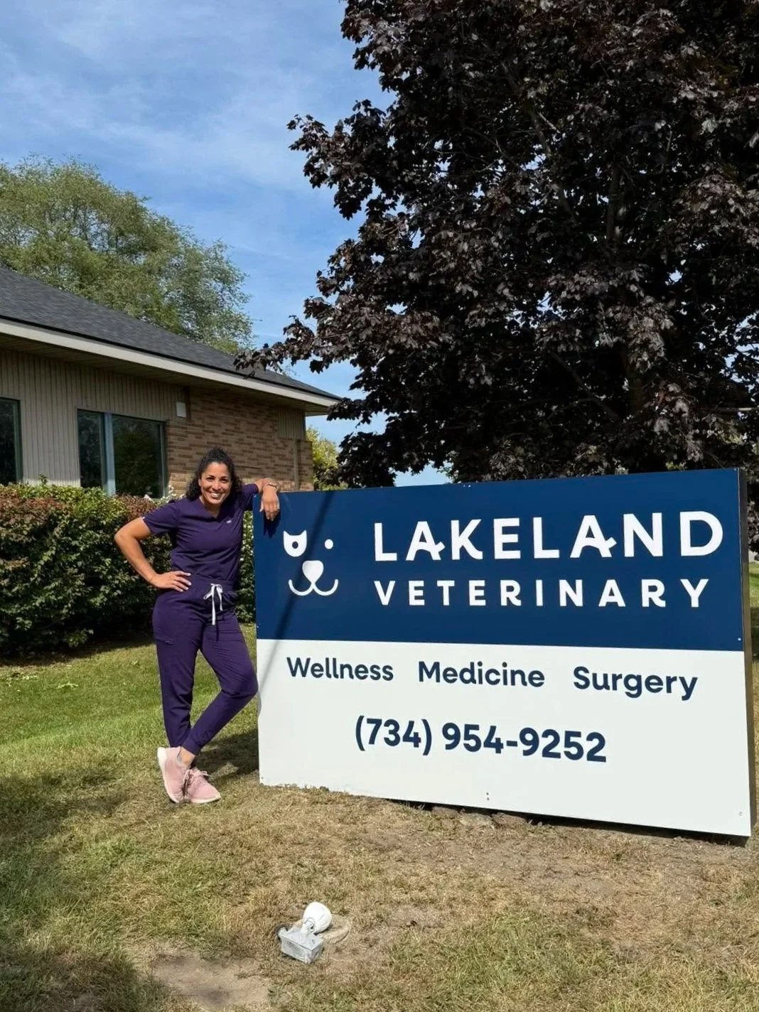 A woman in purple scrubs standing next to a sign for Lakeland Veterinary, which offers wellness, medicine, and surgery services, with a contact phone number. She is outdoors, smiling, with a building, trees, and a blue sky in the background.