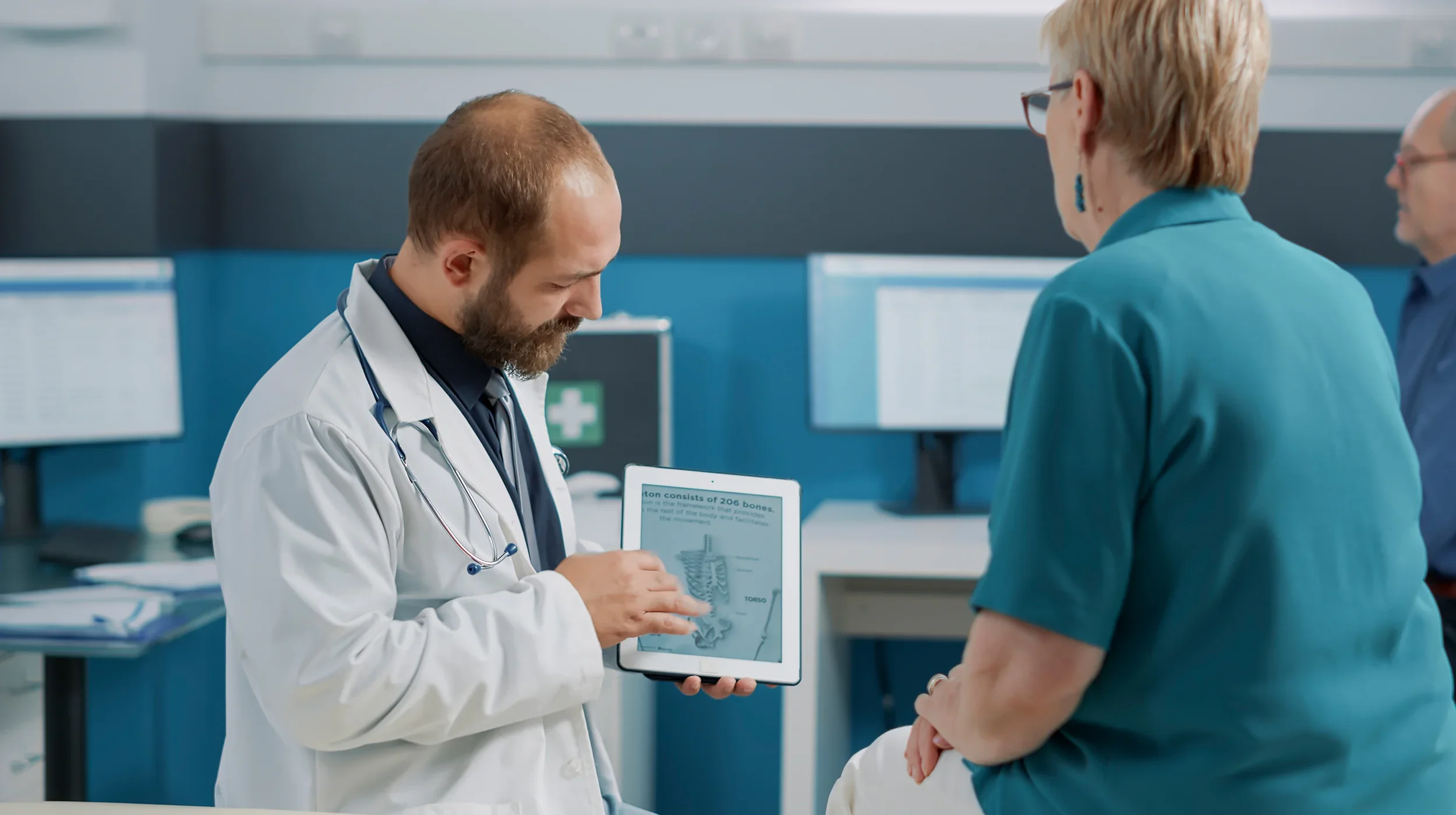 A doctor consulting with an elderly male patient in a medical office, using a tablet device.