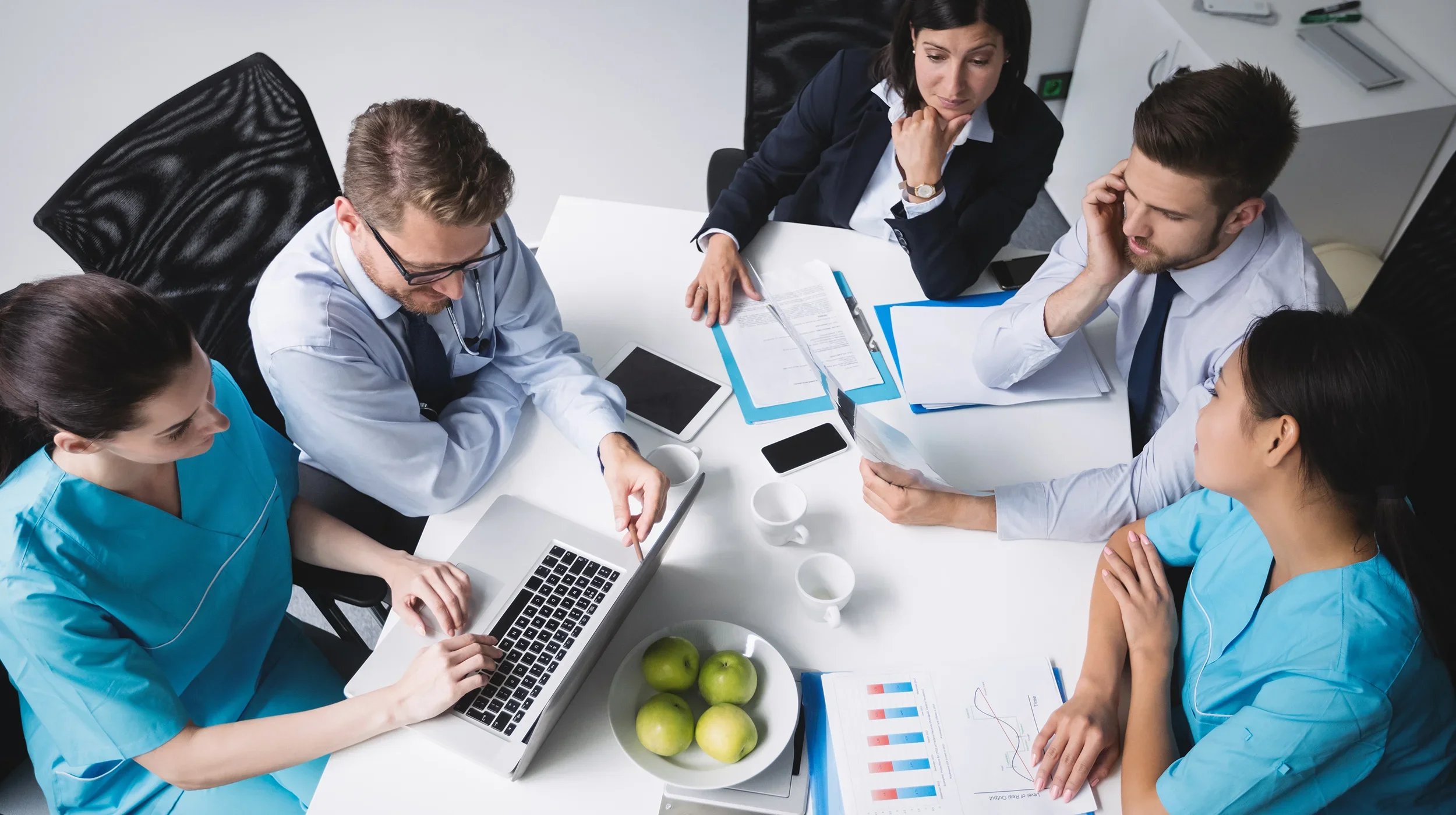 Top-down view of six people working together at a wooden conference table with laptops, tablets, notebooks, pens, and cups of coffee.