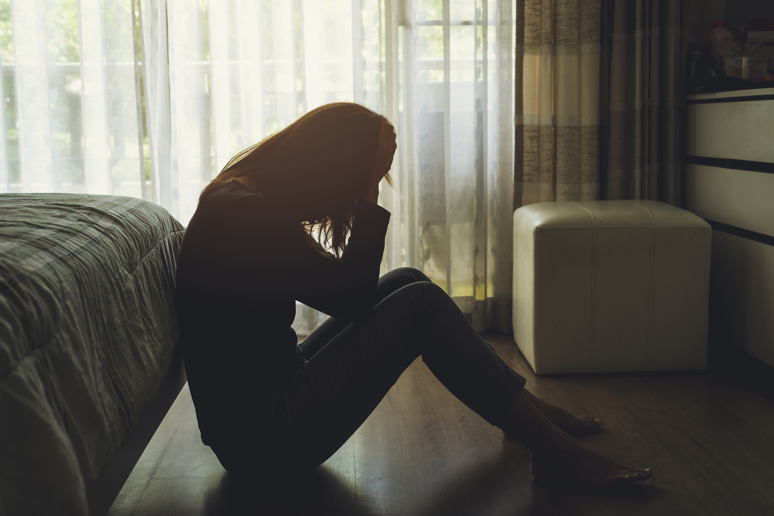 A woman sitting on the floor beside a bed, holding her head in distress, representing depression and emotional struggle.