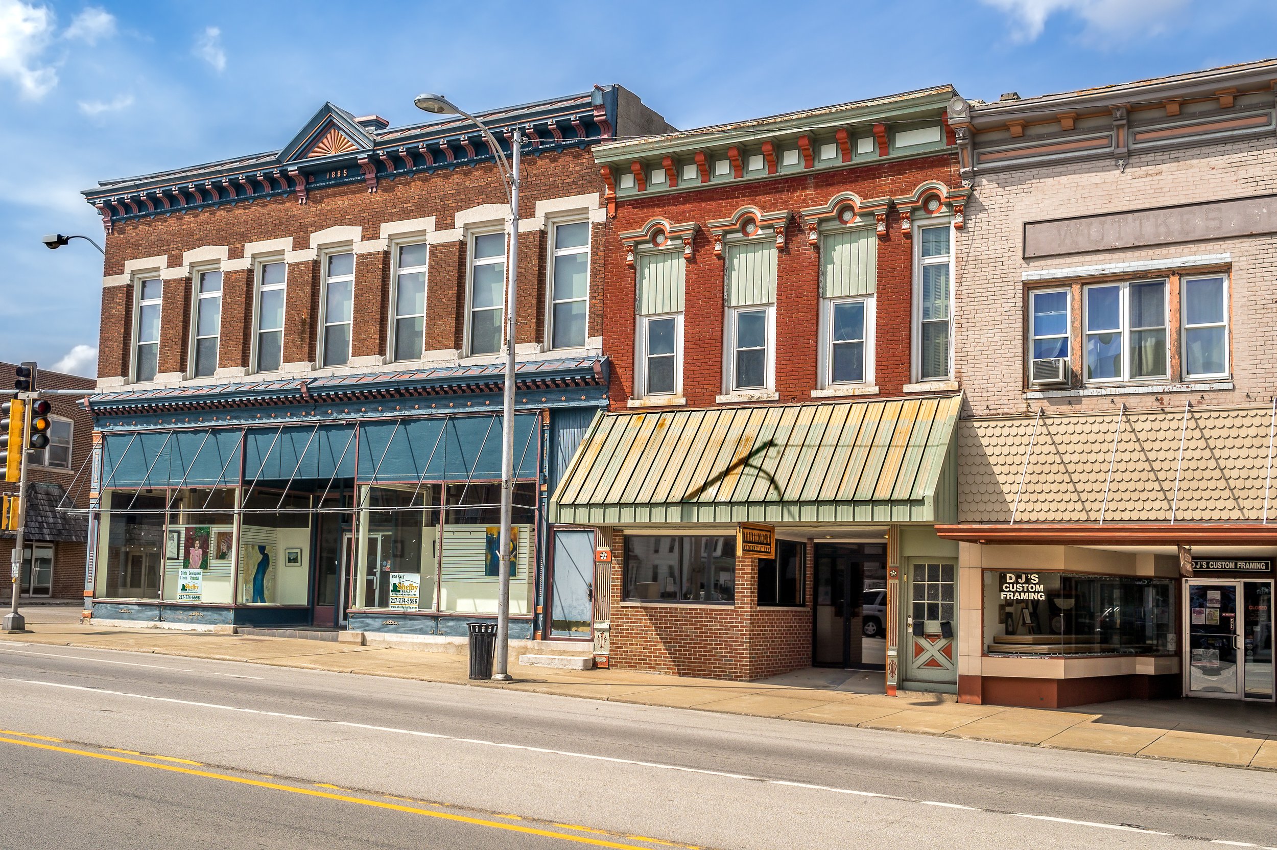 Street view of a row of old brick buildings with commercial storefronts, including a storefront with large windows, a green awning, and the storefront sign 'DJ'S CUSTOM FRAMING.' A traffic light and street lamp are visible, and the sky is partly cloudy.