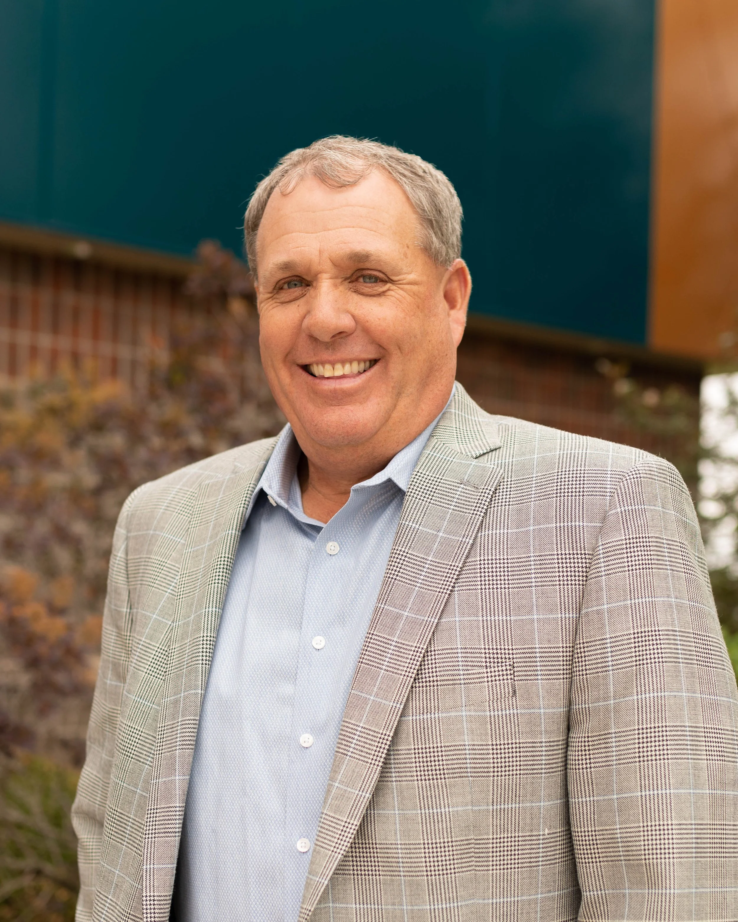 A smiling middle-aged man with light-colored hair, wearing a light blue button-up shirt and a light gray checked blazer, standing outdoors with a blurred background of a brick wall and trees.