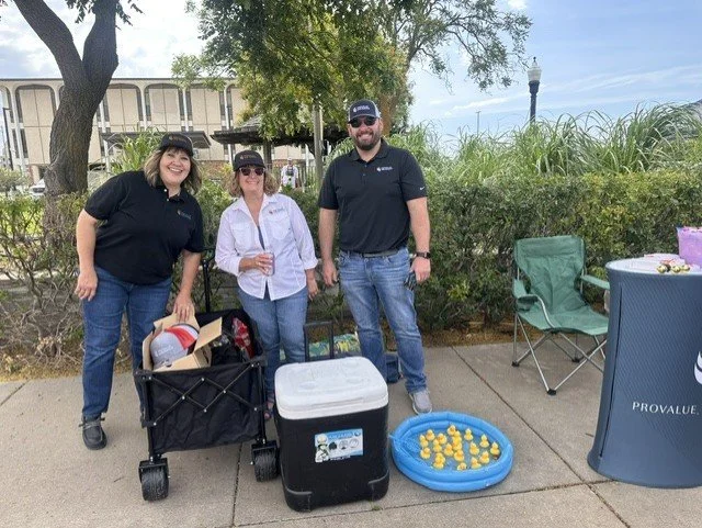 Three people standing outdoors next to a small kiddie pool filled with rubber duckies, with cooler and bags nearby.