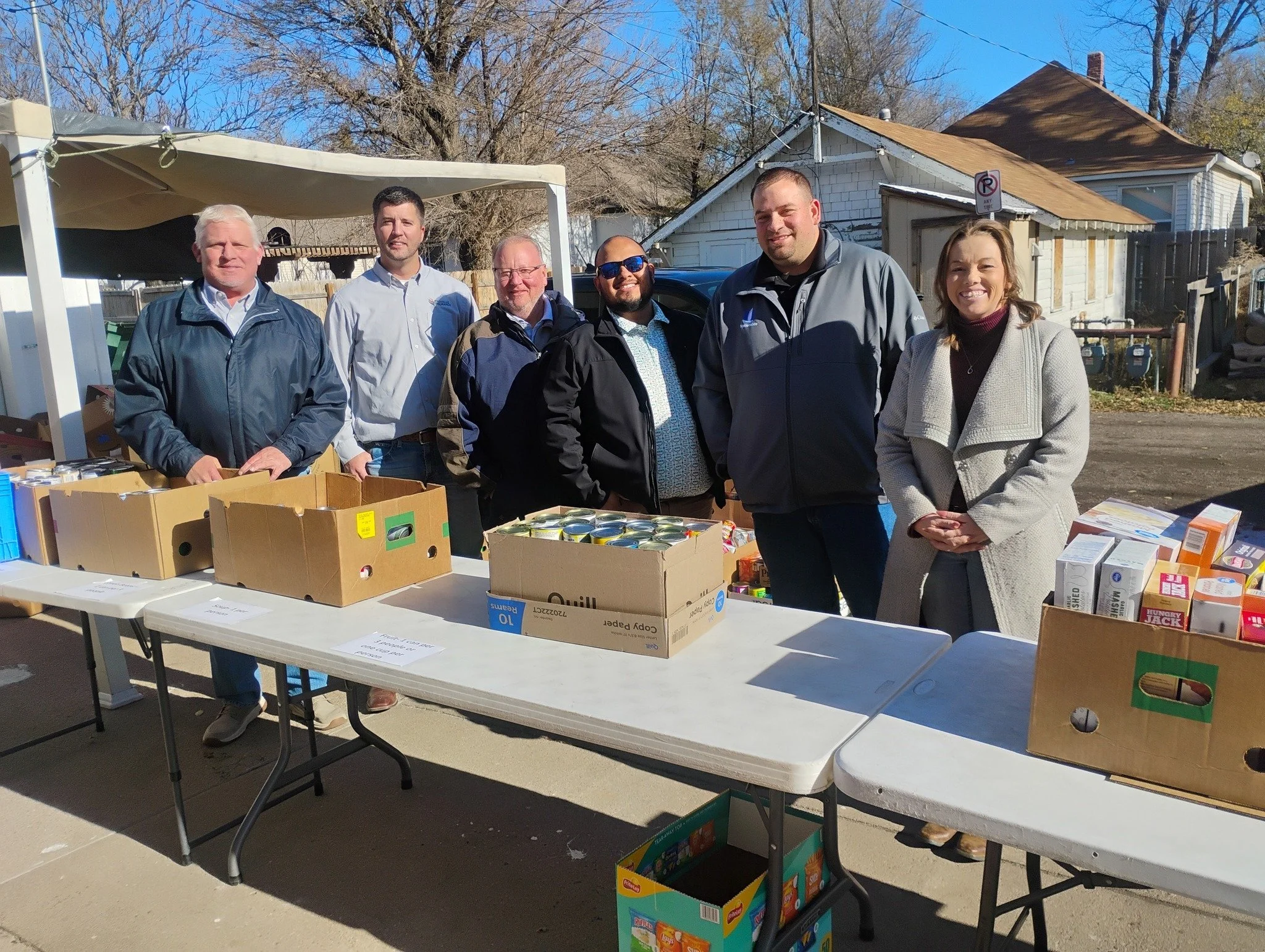 A group of six people standing behind tables with boxes of canned goods and food items, outdoors on a sunny day, in front of residential houses.