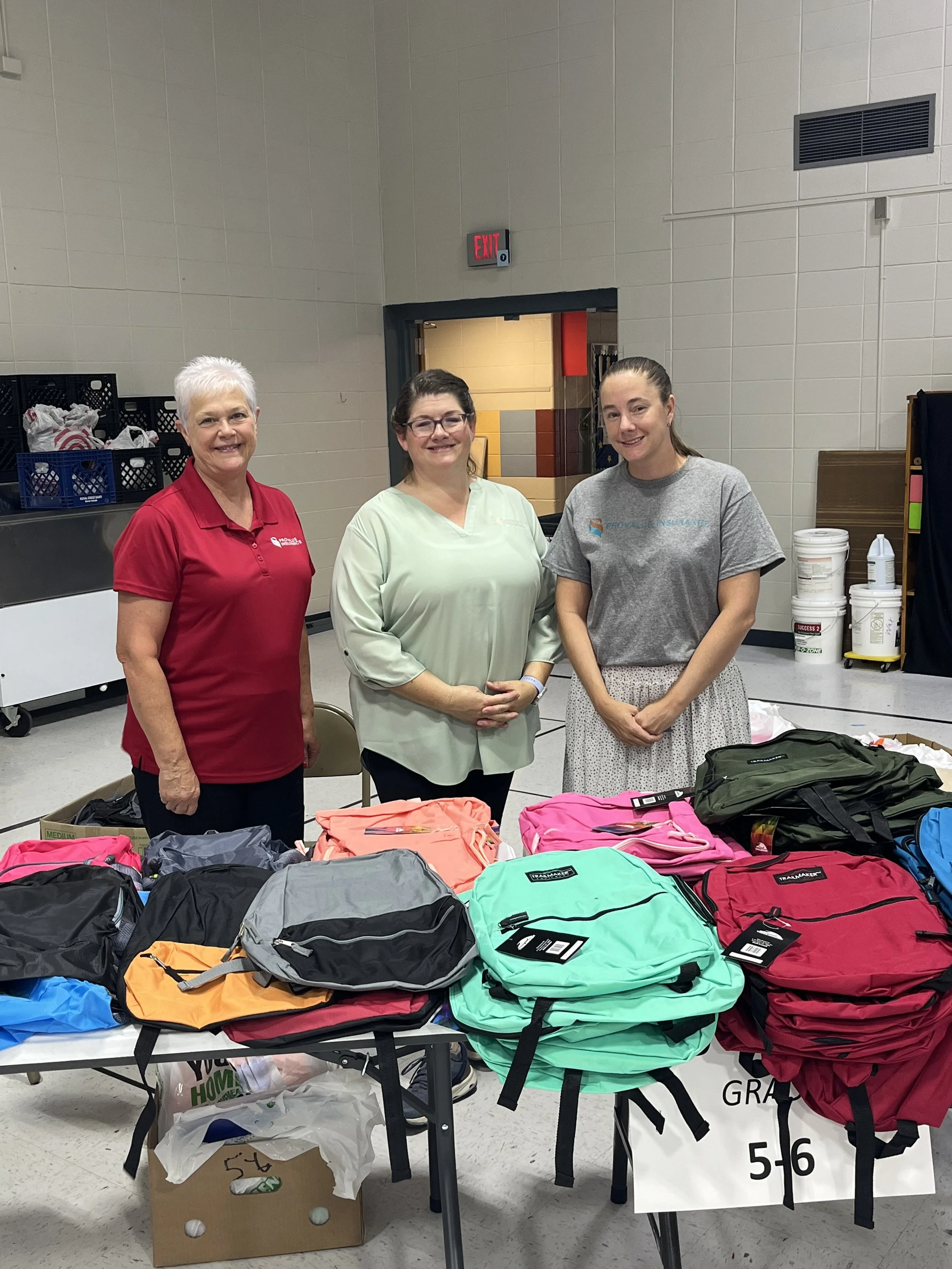 Three women smiling behind tables filled with colorful backpacks and supplies, in a large indoor gathering space.