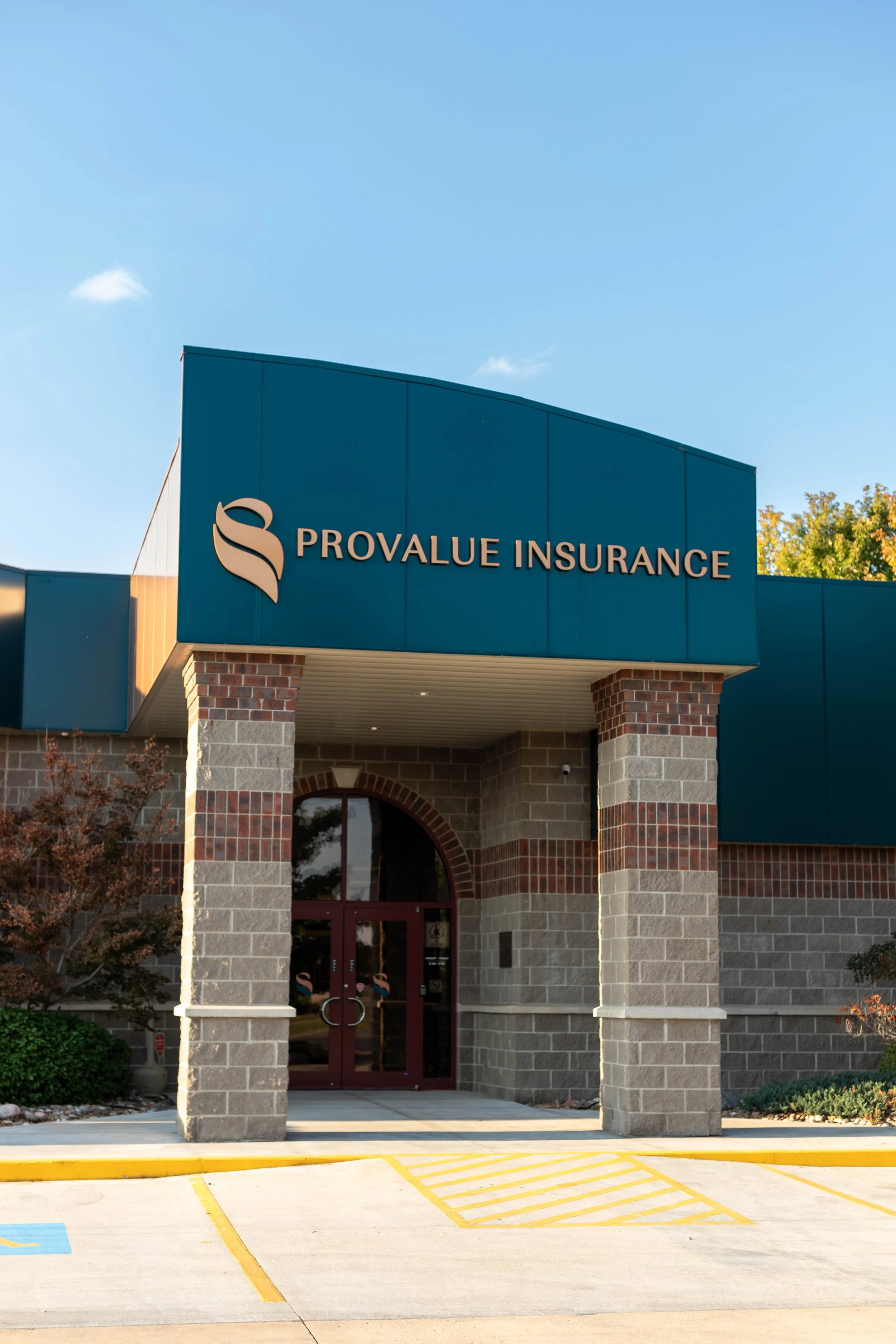 Exterior of ProValue Insurance building with brick columns and blue signage, parking lot with painted lines in the foreground, and trees with fall foliage in the background.