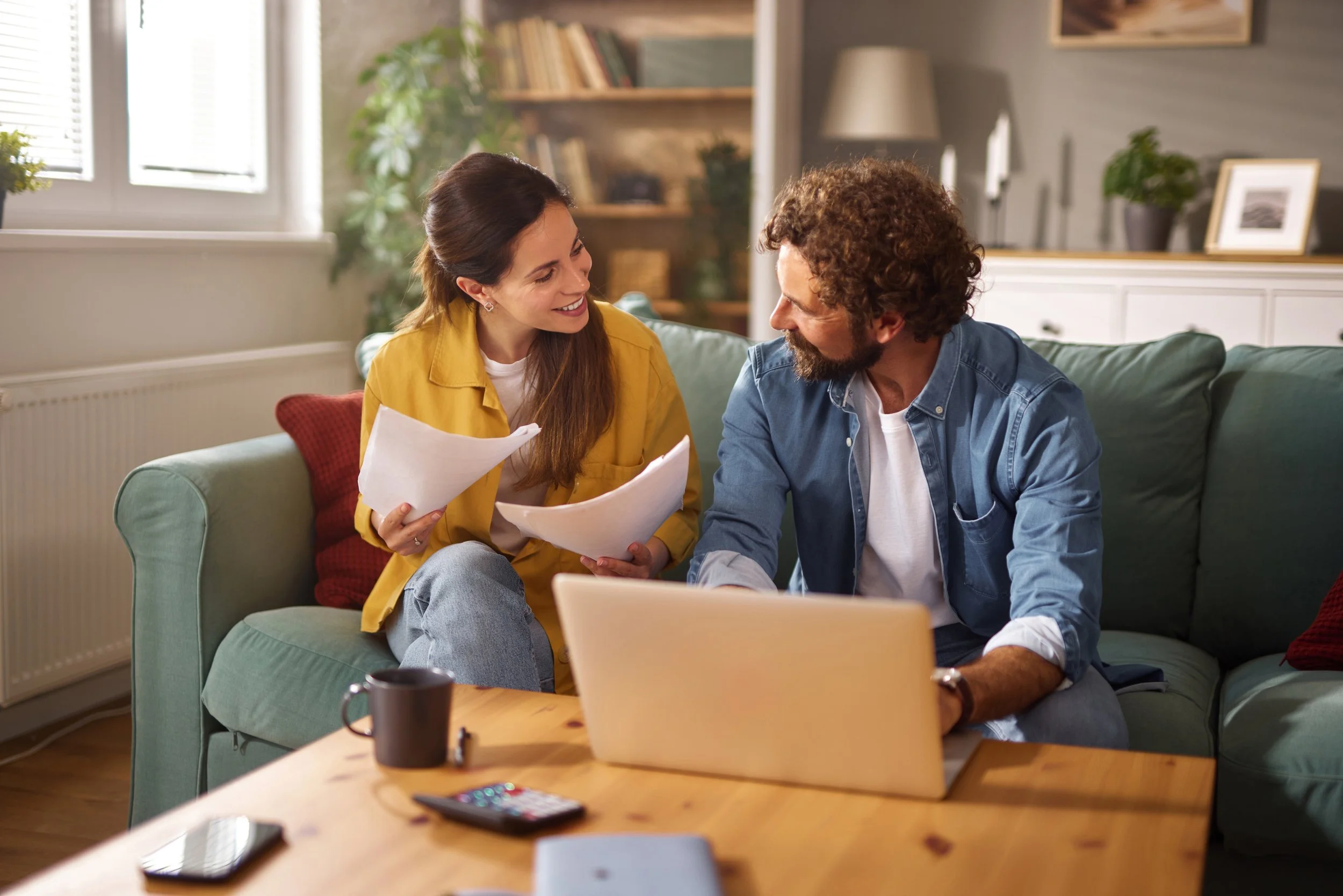A woman in a mustard yellow jacket holding papers and smiling while talking to a man with curly hair and beard, wearing a blue denim shirt, sitting on a couch in a cozy living room with a laptop on the table in front of them.