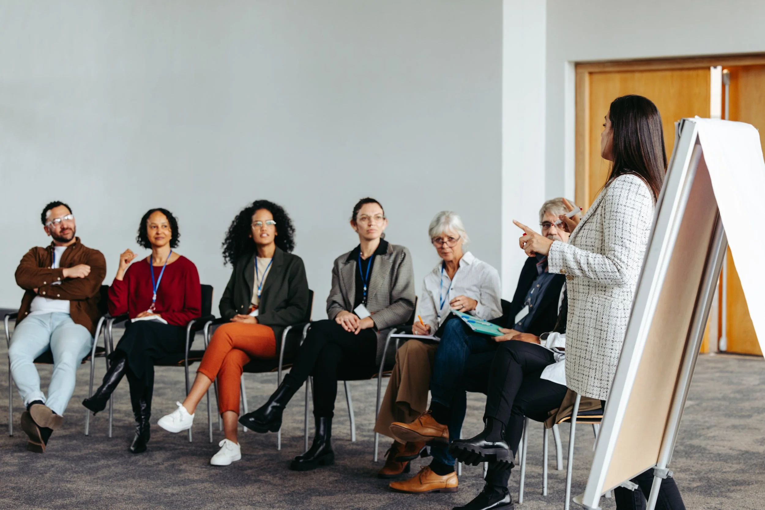A diverse group of people seated in a line, listening to a woman speaker who is standing with a notepad, in a conference room or classroom setting.