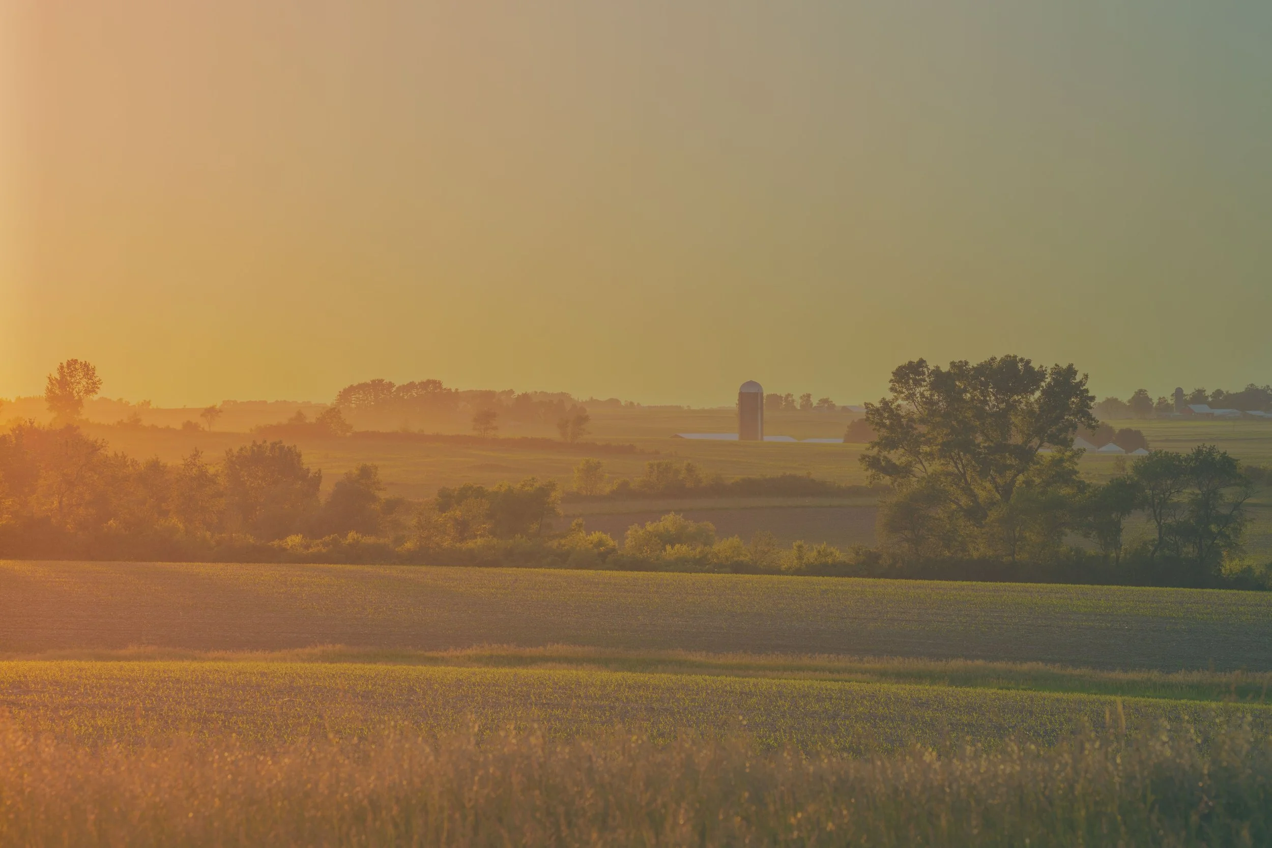 A rural landscape at sunset with fields, trees, and farm buildings in the distance.