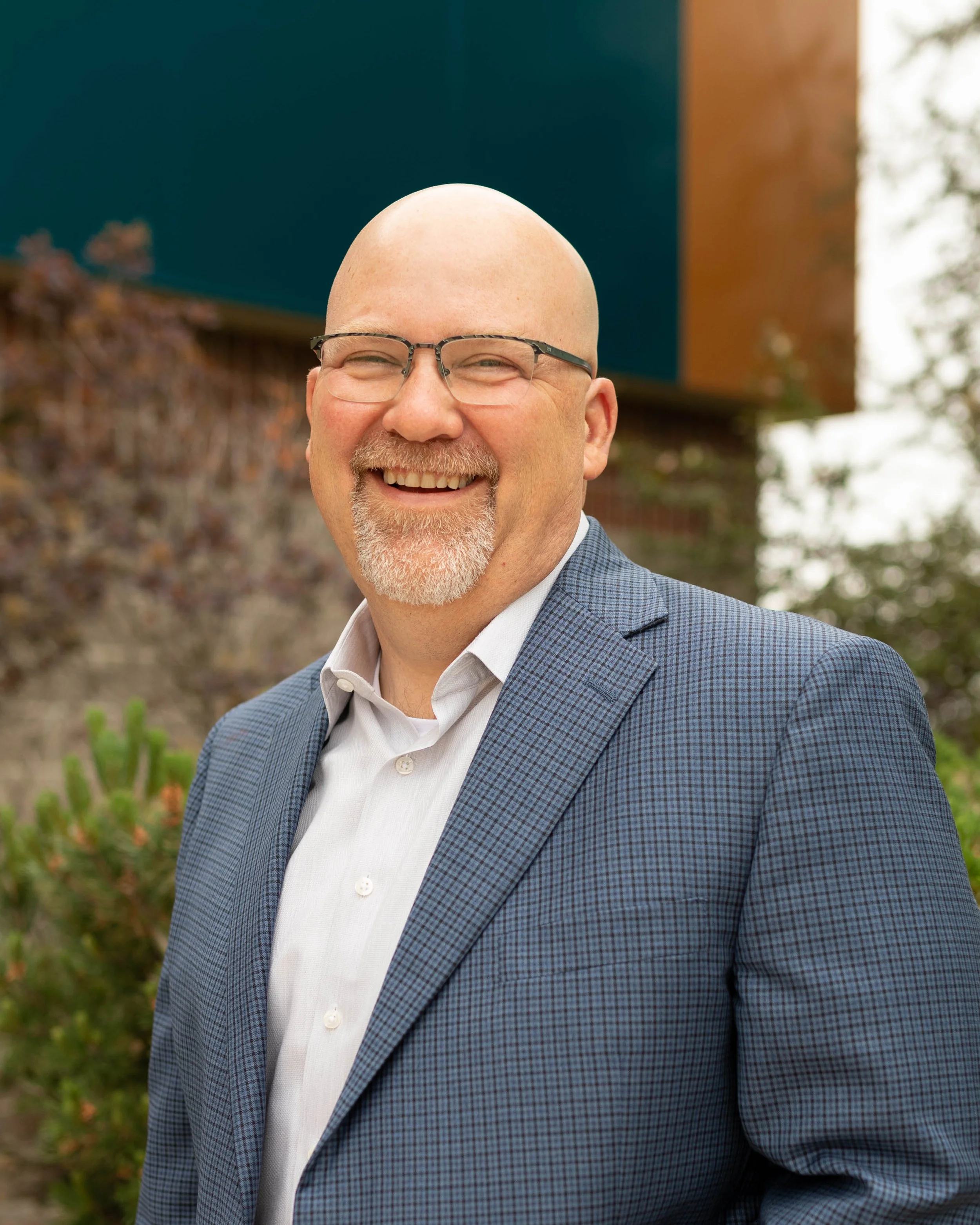 A smiling bald man with glasses, wearing a checkered blue blazer and white shirt, standing outdoors with a blurred background of trees and a building.
