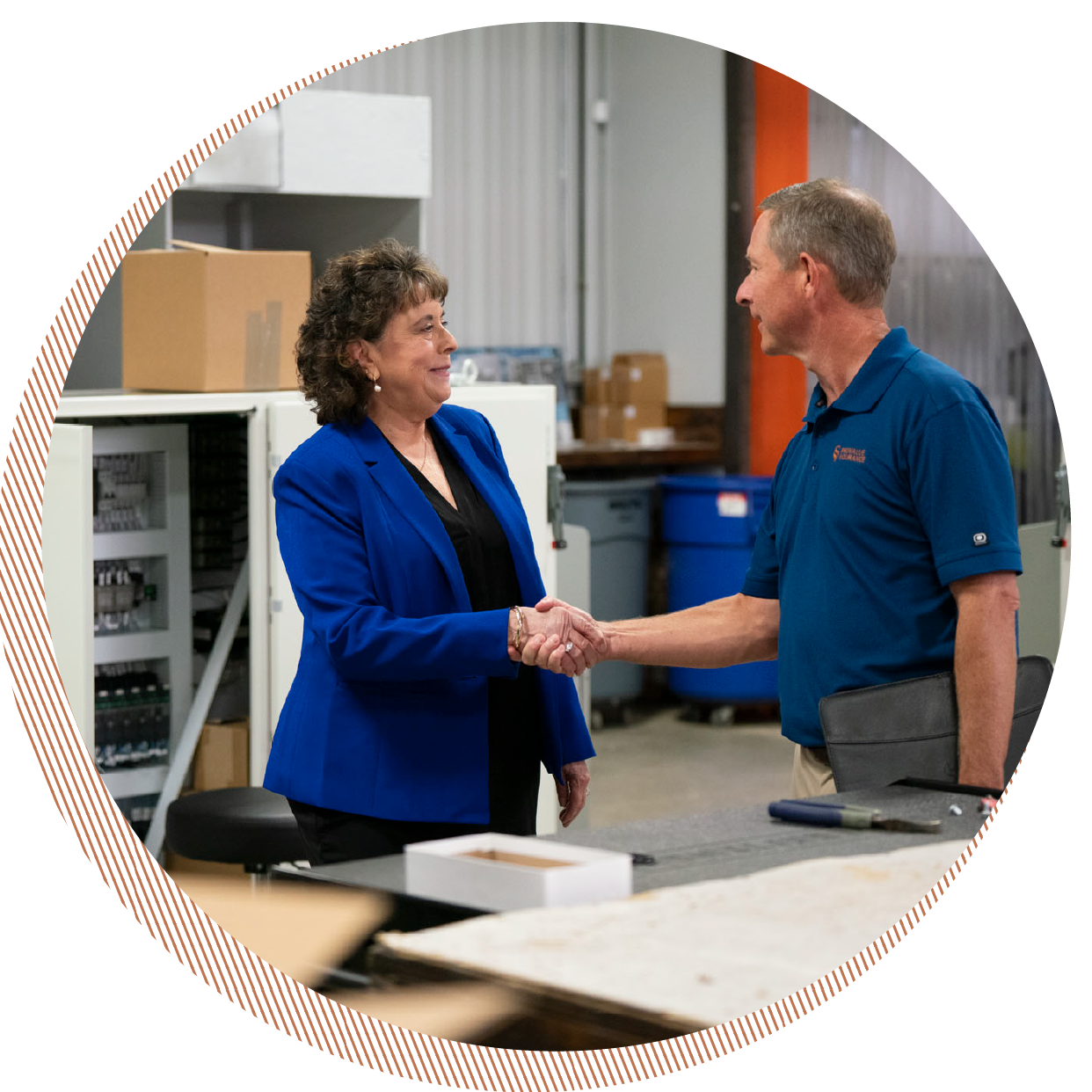 A woman and a man shaking hands in an industrial or warehouse setting, both smiling. The woman is wearing a blue blazer and the man is wearing a blue polo shirt.