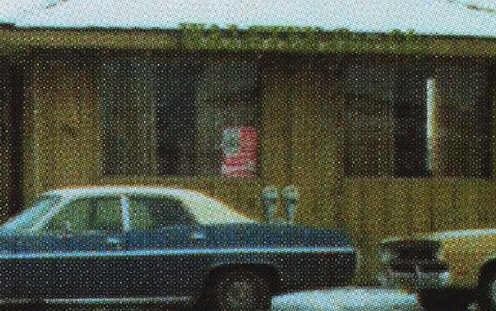 A vintage black car parked in front of a wooden building with two windows and a door in an outdoor setting.