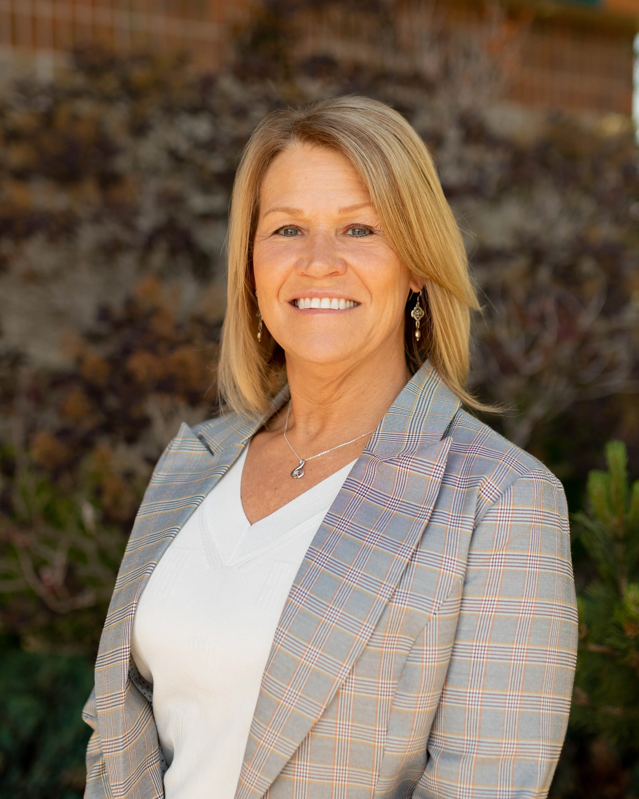 A woman with blonde hair wearing a checked blazer, a white top, a necklace, and earrings, smiling outdoors with a blurred background of brown and green foliage.