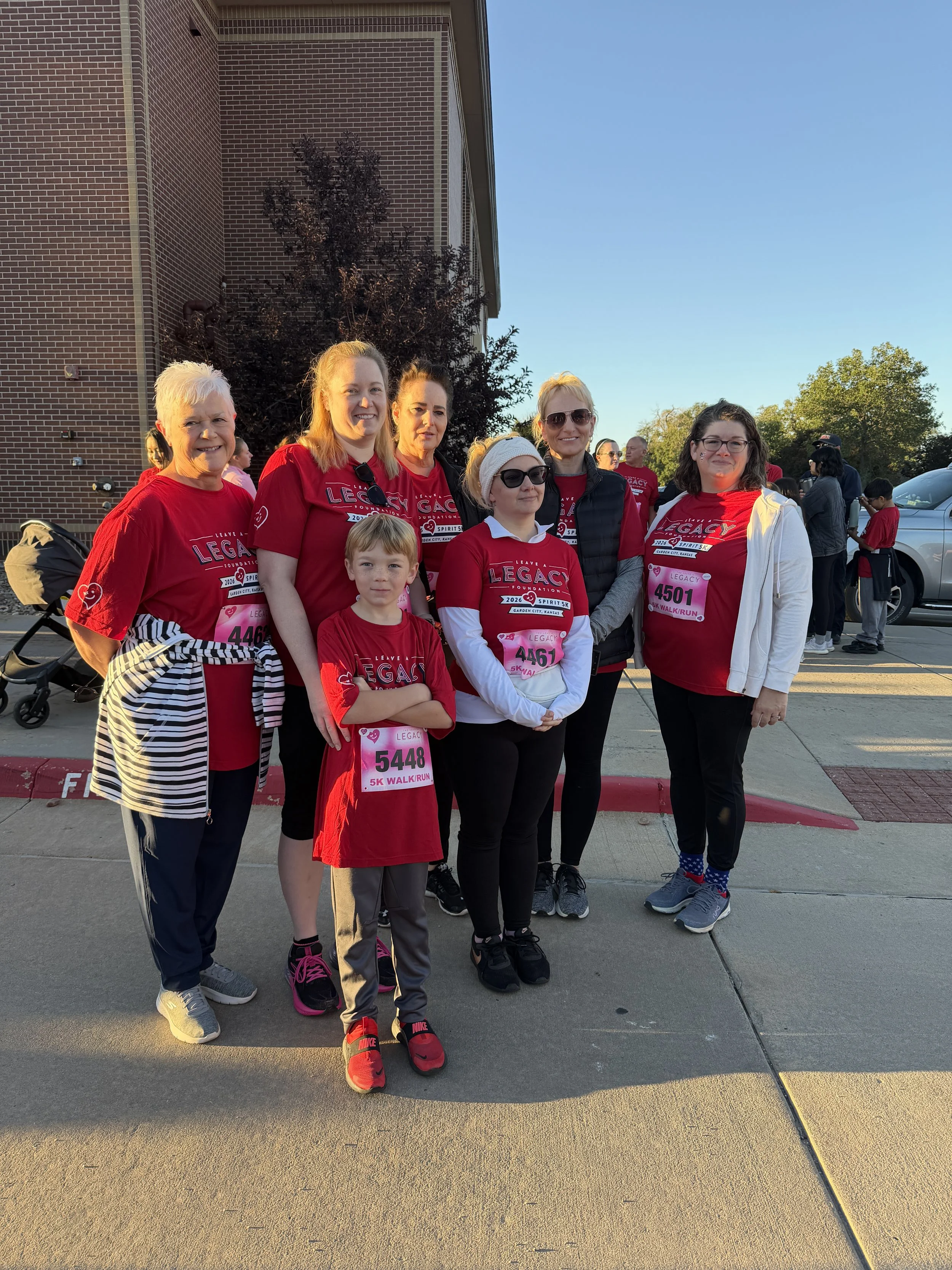 Group of people, including children and adults, gathered outdoors for a walk/run event, wearing matching red shirts with event details, numbered bibs, and casual athletic attire, standing on a sidewalk with a brick building and trees in the backgroun