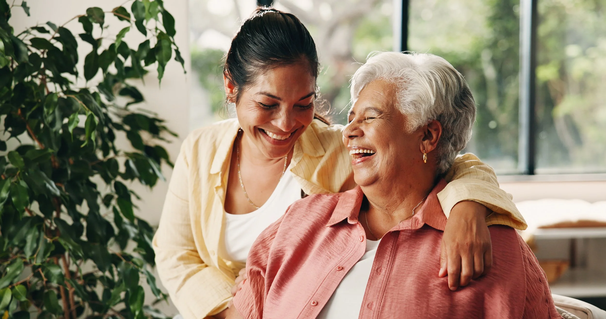 A young woman hugging and smiling at an elderly woman, both indoors with large windows and green plants in the background.