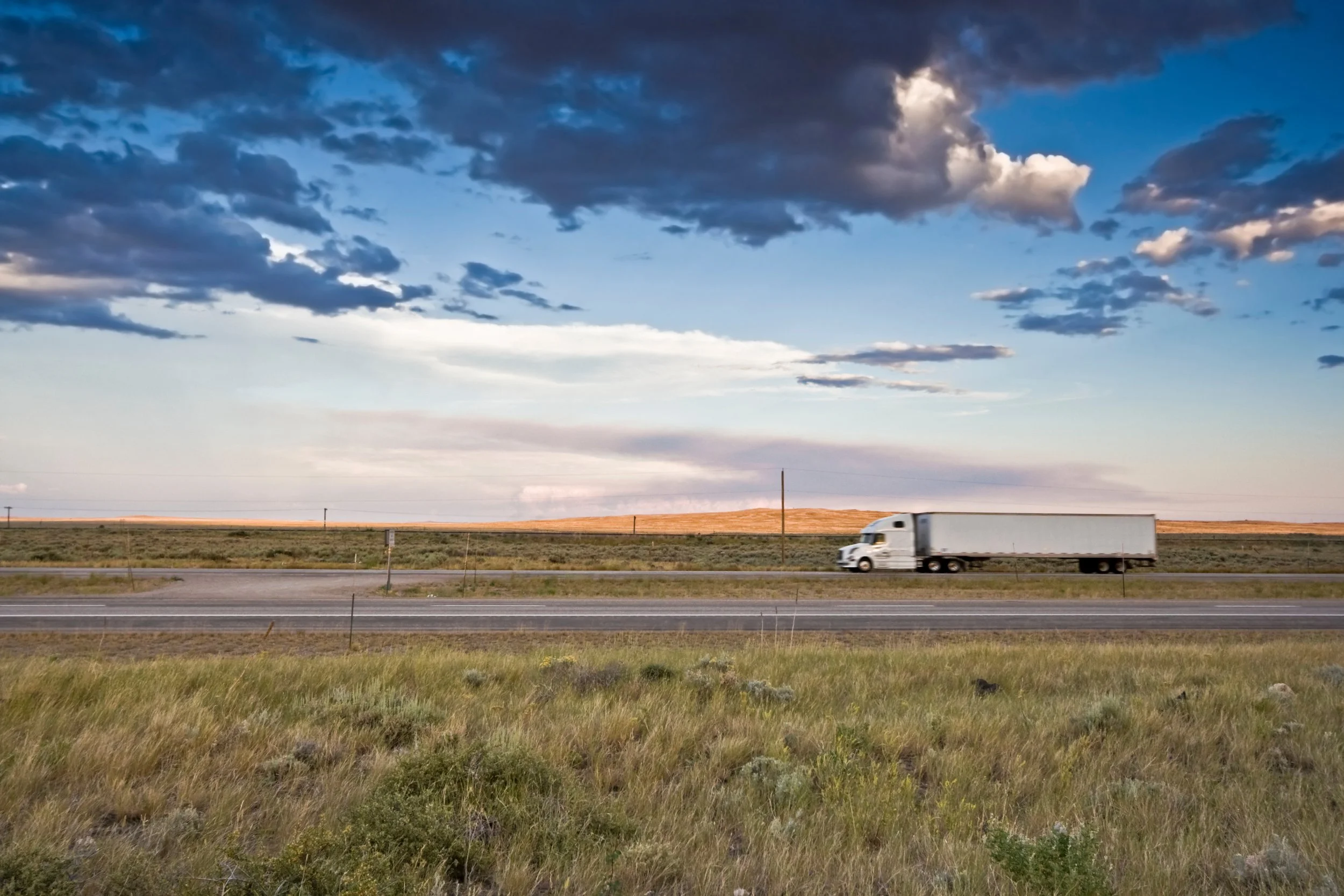 Open highway with a moving semi-truck and a cloudy sky over grassy plains.