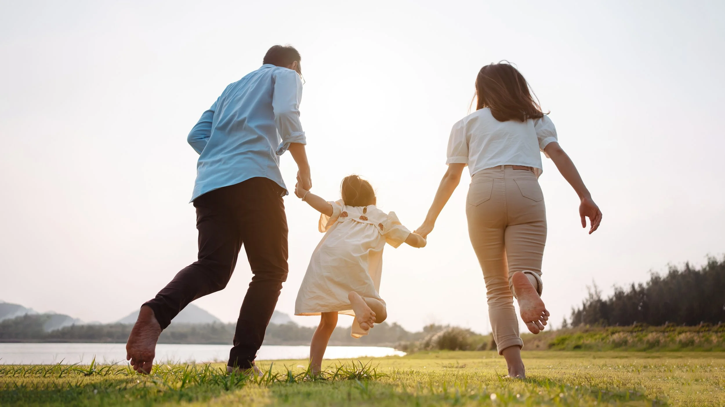 A family of four holding hands and walking together outdoors at sunset, with a body of water and trees in the background.